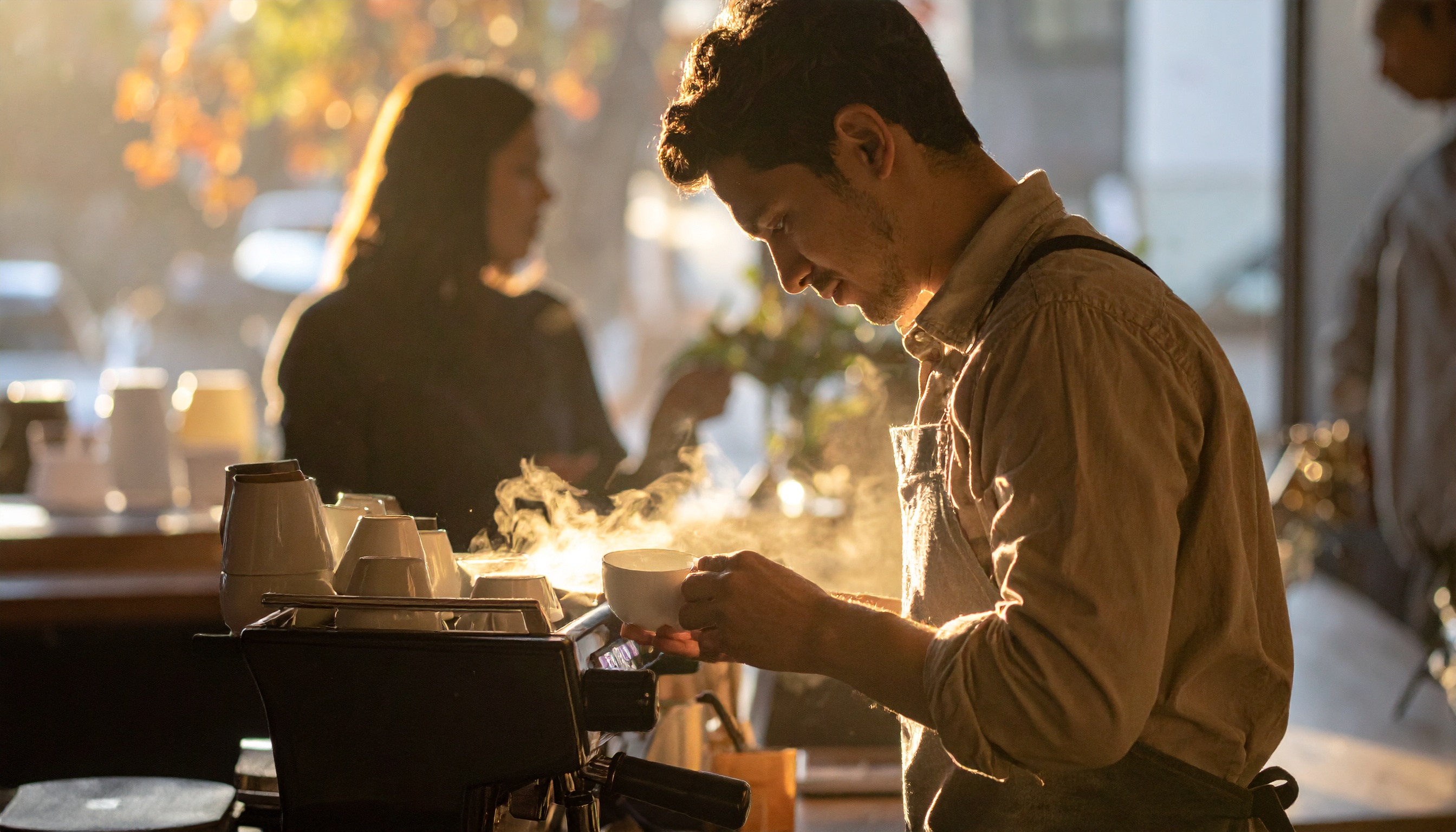 Barista focado prepara uma xícara de café em um ambiente iluminado pelo sol da manhã, destacando vapor do café sob luz dourada suave. Cena transmite tranquilidade em um café movimentado, com fundo desfocado mostrando silhuetas de clientes interagindo.