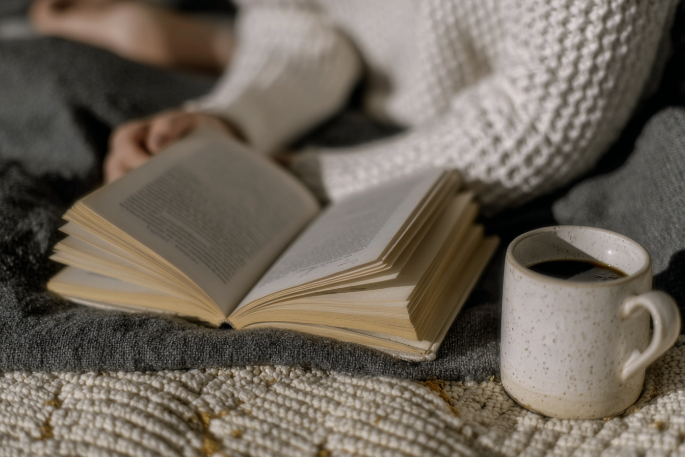 A imagem apresenta uma cena aconchegante com um livro aberto sobre uma superfície de tecido cinza, ao lado de uma caneca de cerâmica cheia de café. A pessoa, vestida com um suéter de tricô branco, segura o livro, sugerindo um momento relaxante de leitura. A iluminação suave e difusa cria uma atmosfera de tranquilidade e conforto, com foco seletivo destacando os objetos principais em primeiro plano.