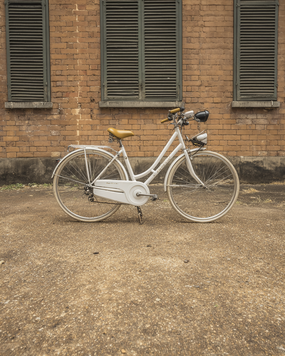 Bicicleta branca com detalhes em couro marrom claro, estacionada em frente a uma parede de tijolos antigos com janelas de venezianas verdes. A composição é centralizada, destacando o contraste entre a modernidade da bicicleta e o estilo rústico da parede. A iluminação suave cria uma atmosfera nostálgica, enquanto a textura do chão adiciona profundidade visual à cena.