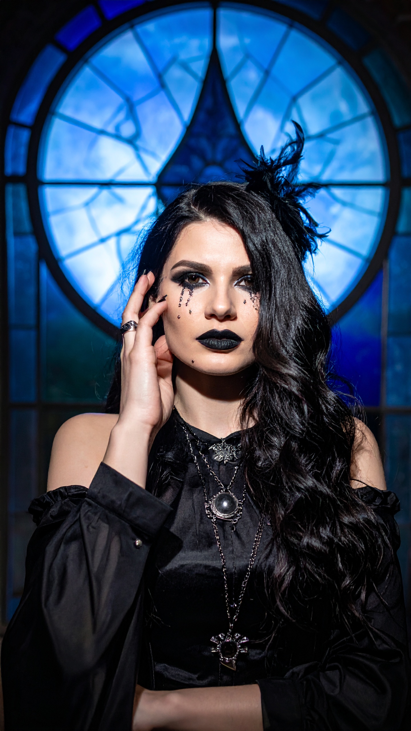 Mulher em estilo gótico posando em frente a um vitral azul impressionante. Ideal para eventos temáticos ou sessões de fotos artísticas. Detalhes como maquiagem escura e acessórios carregados dão um toque dramático. Perfeito para festas de Halloween ou encontros culturais alternativos. O visual destaca-se pela combinação de luz e sombra, criando um efeito visual cativante.