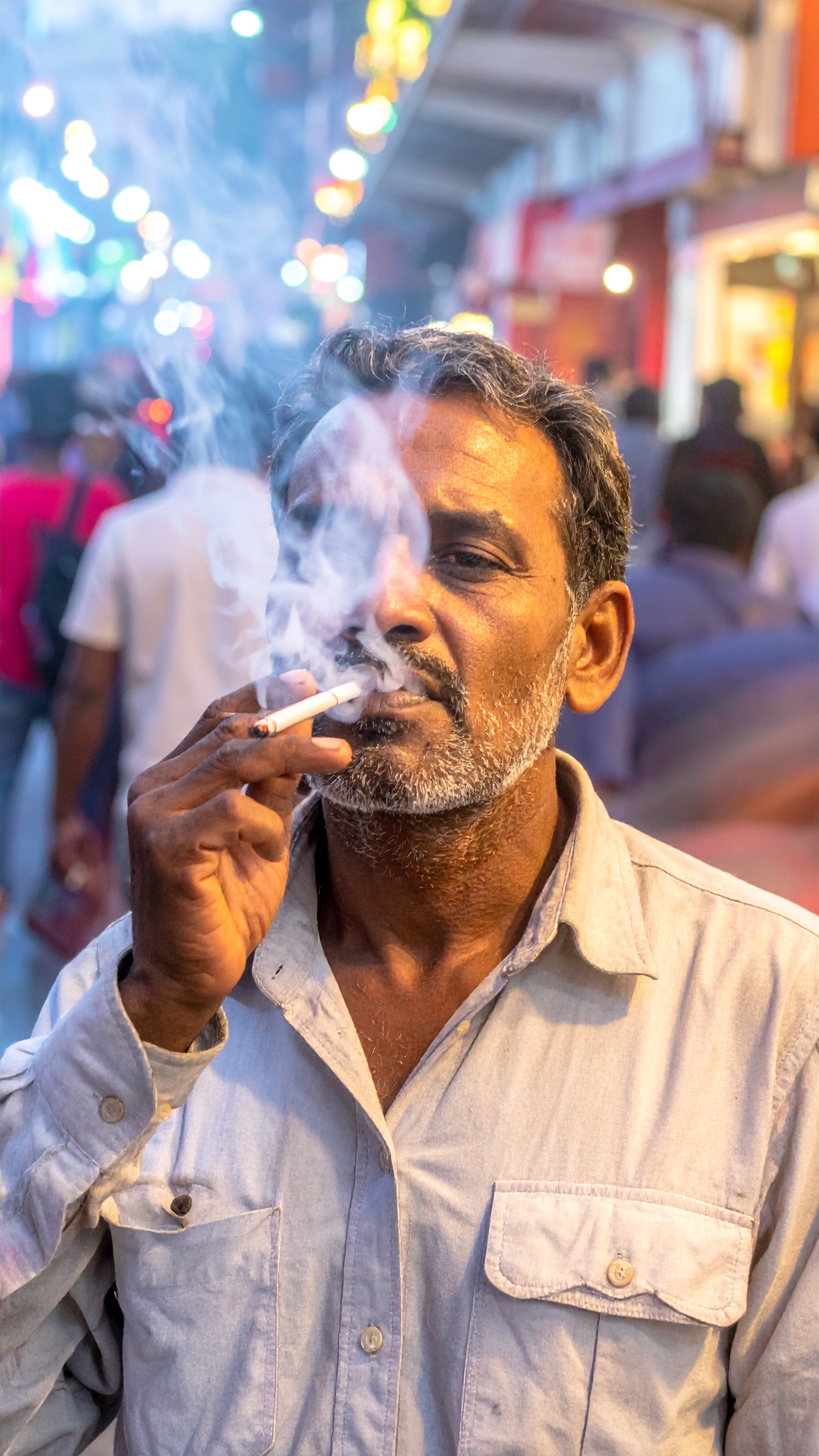 A man smoking a cigarette stands in a bustling street, with vibrant lights in the background
