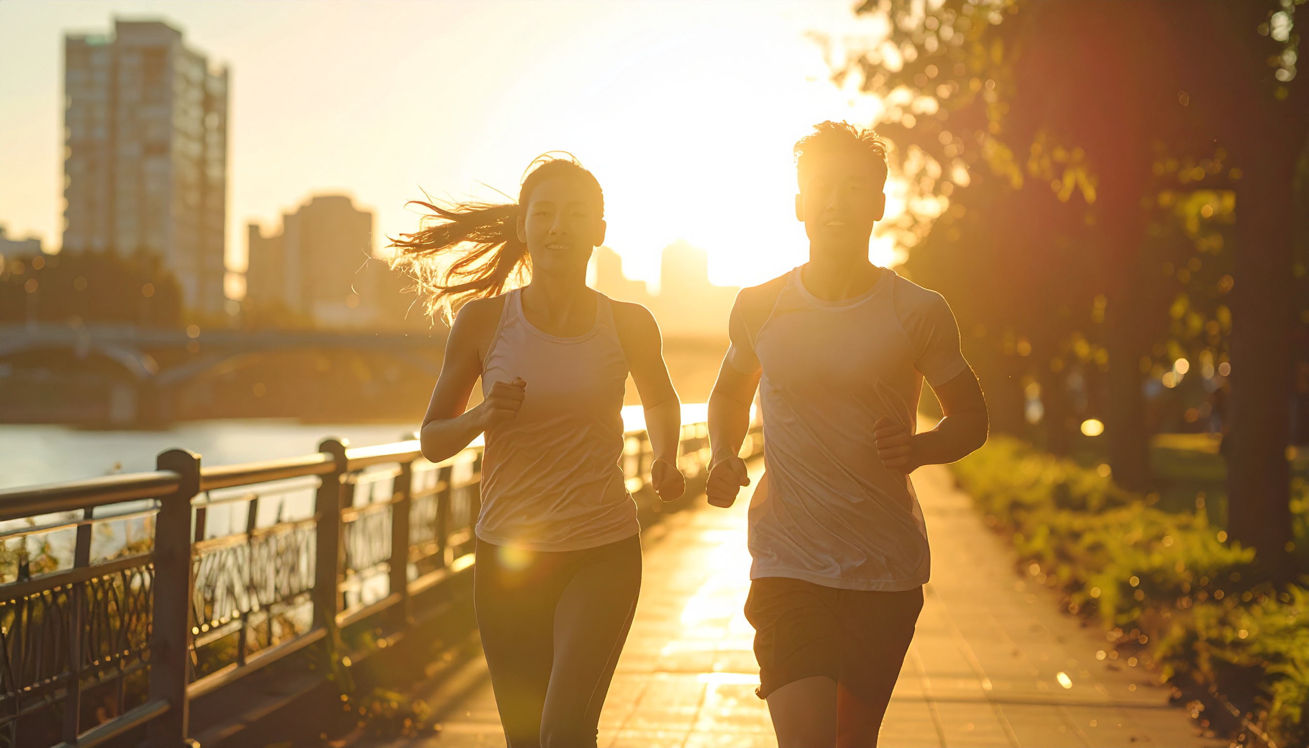 Duas pessoas correm juntas em uma calçada ao longo de um rio, iluminadas por um pôr do sol vibrante. A cena é composta com um fundo urbano desfocado, onde prédios e uma ponte são visíveis. A luz dourada do sol cria um efeito de silhueta, enfatizando o movimento e a energia dos corredores contra o céu brilhante. A composição enfatiza dinamismo e vitalidade, com um ângulo que captura a sensação de liberdade e bem-estar.