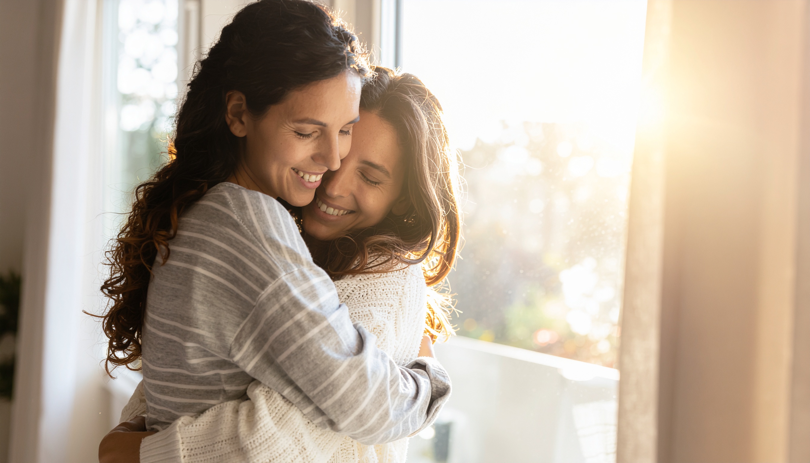 Duas mulheres abraçando-se carinhosamente à luz do sol em uma sala aconchegante, transmitindo afeto e alegria em um ambiente doméstico acolhedor.
