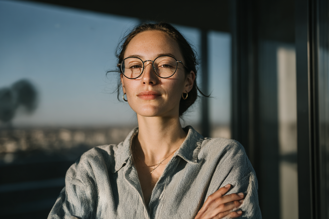 Retrato de uma pessoa com expressão confiante, iluminada pela luz suave do sol, usando óculos redondos e camisa texturizada em tons neutros.