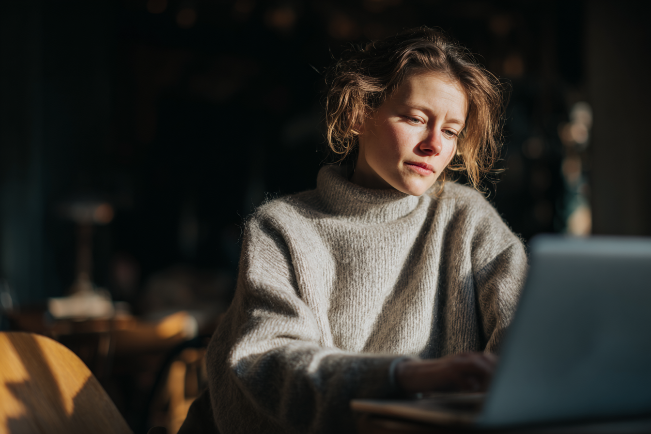 Mulher em suéter cinza, concentrada em laptop em ambiente interno. Luz natural suave ilumina o rosto, criando um ambiente acolhedor e introspectivo, com foco suave e fundo desfocado.