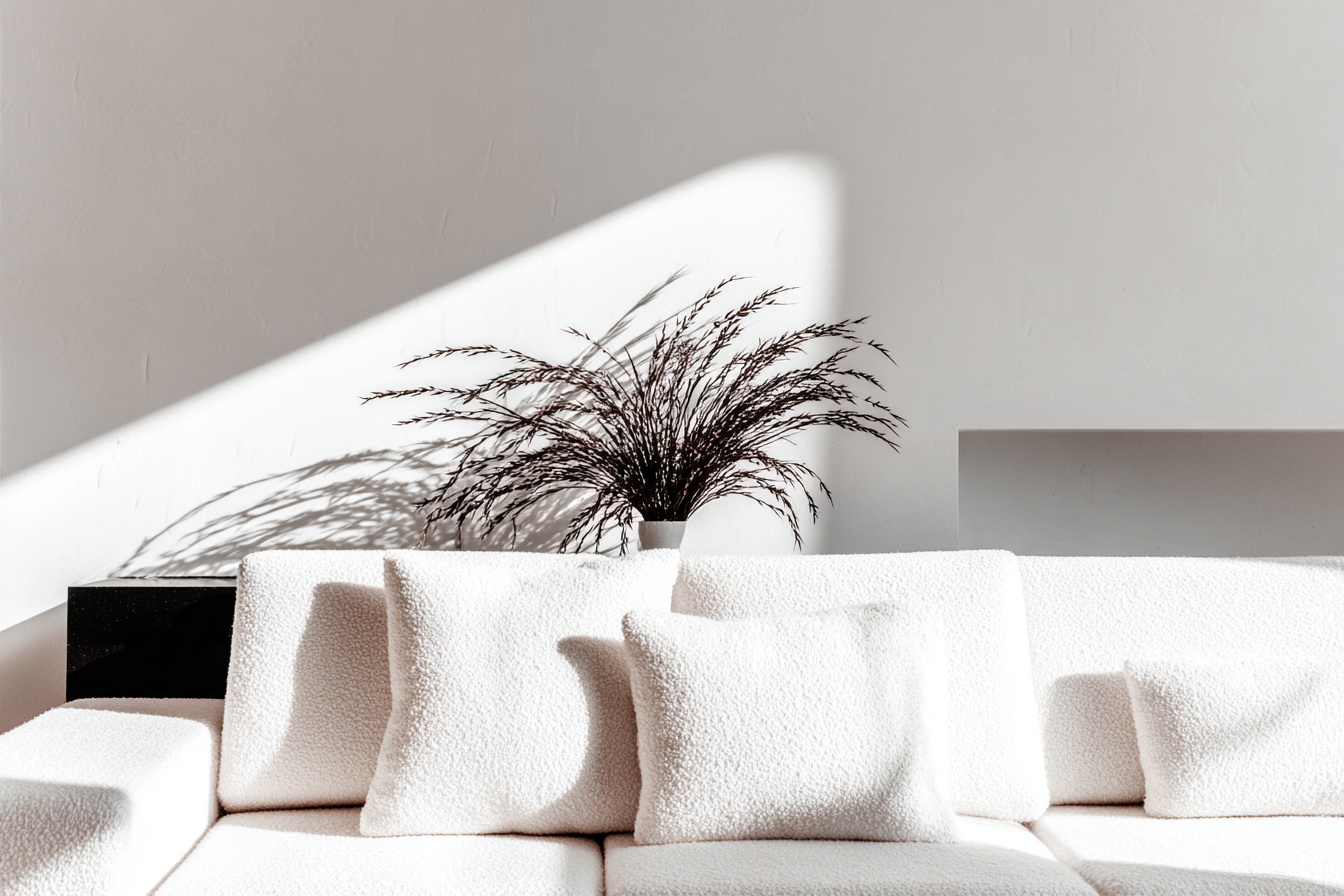 Sofá moderno com almofadas brancas em ambiente minimalista iluminado por luz natural, destacando um vaso decorativo com ramo seco sobre superfície clara, evocando tranquilidade e sofisticação.