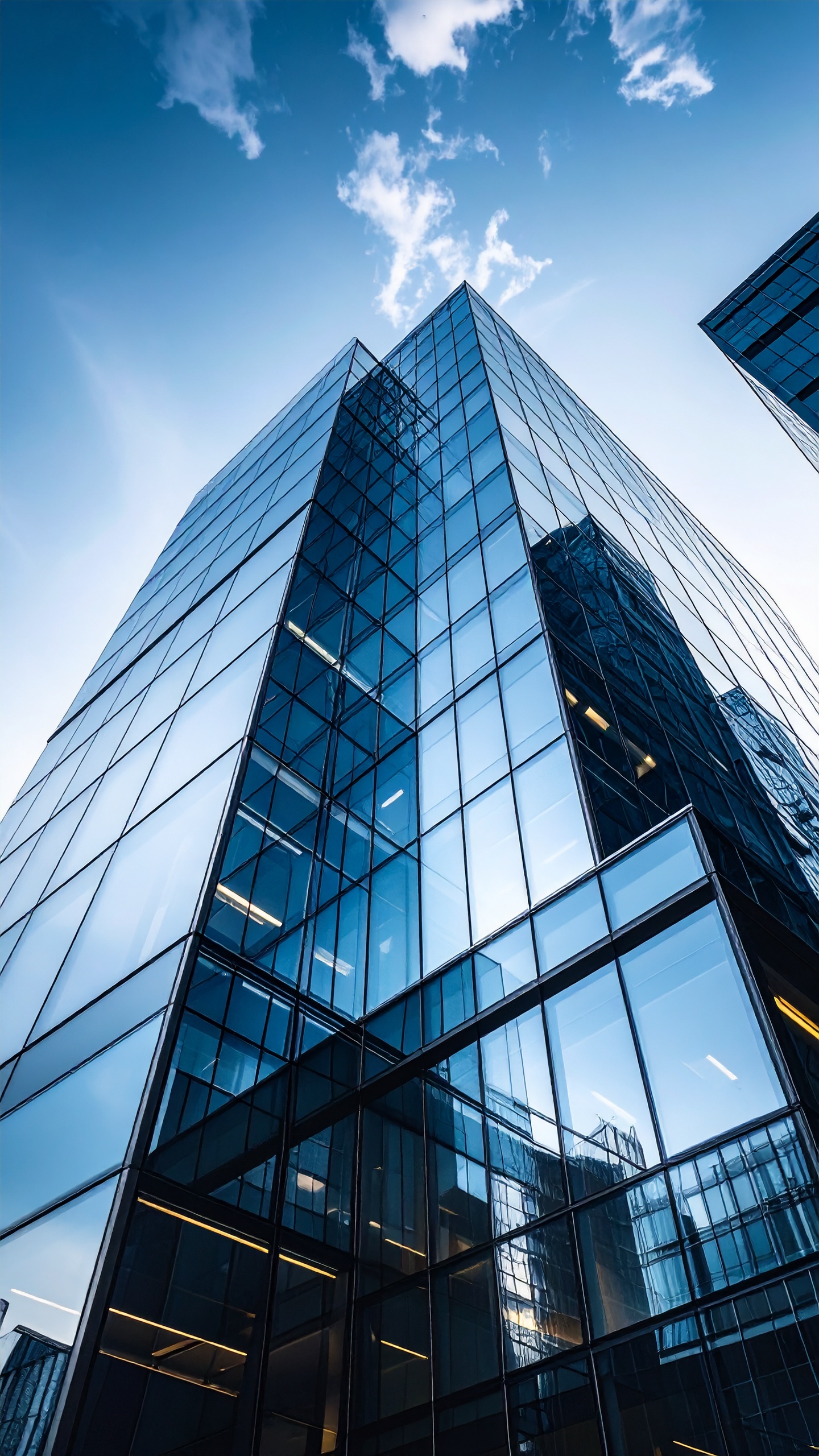 Imagem de um arranha-céu moderno com fachada de vidro refletindo o céu azul e nuvens, capturada de um ângulo baixo que acentua a altura e verticalidade da estrutura. A iluminação natural destaca as linhas geométricas e cria um contraste entre a arquitetura e o céu, conferindo uma atmosfera de modernidade e imponência urbana.