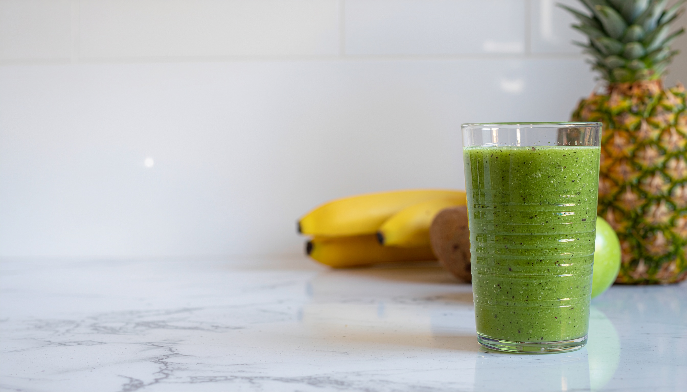 Smoothie verde vibrante em copo de vidro sobre bancada de mármore, com bananas, abacaxi e maçã no fundo, iluminação clara e fundo minimalista.