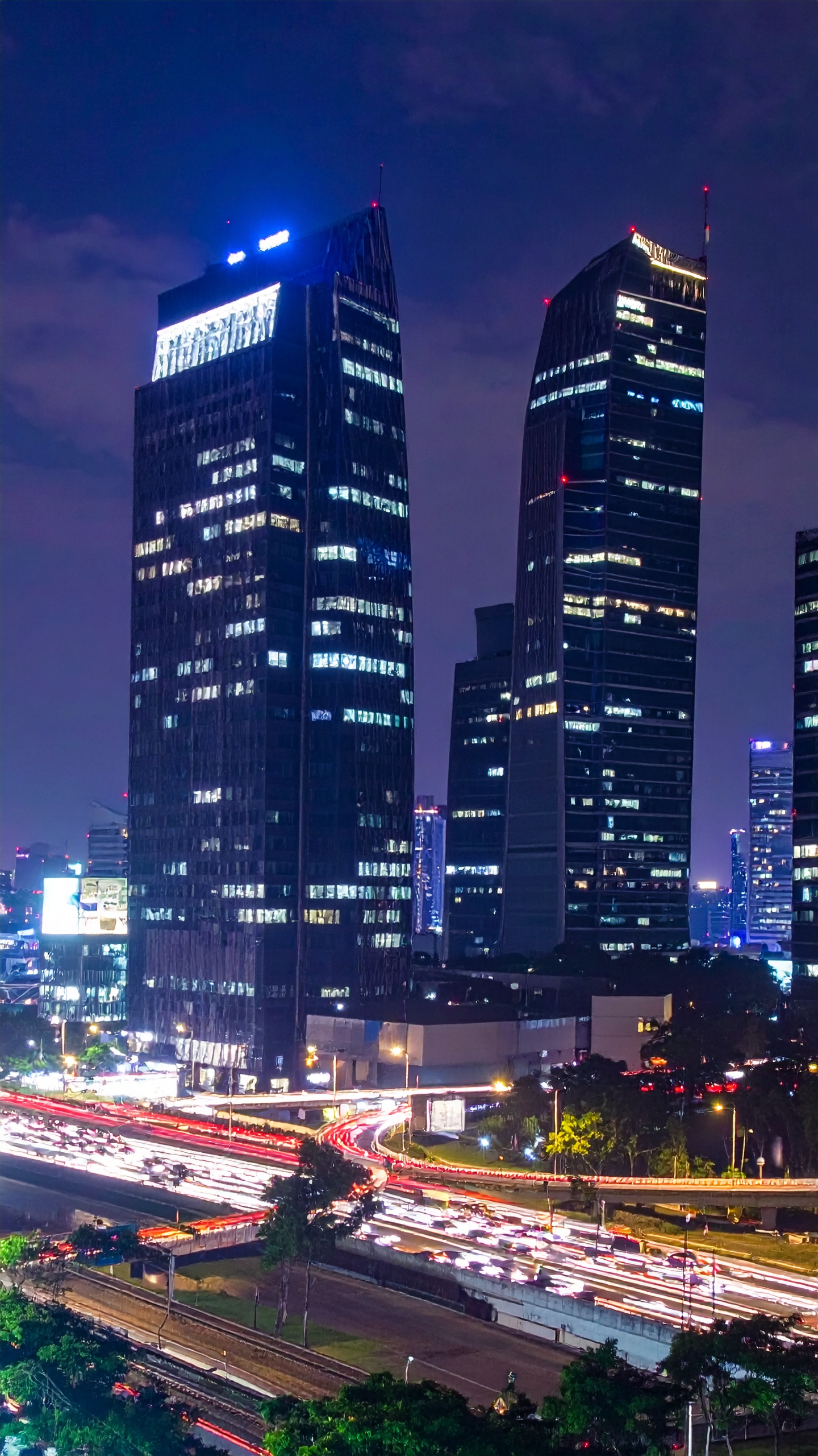 Prédios modernos iluminados à noite sobre uma movimentada avenida com tráfego intenso. Ideal para representar a vida urbana e a modernidade em campanhas publicitárias. As luzes dos edifícios contrastam com o céu escuro, criando um efeito visual impressionante. Perfeito para uso em materiais de marketing que destacam ambientes urbanos e inovação. Um toque de dinamismo e sofisticação que pode ser explorado em várias aplicações visuais.