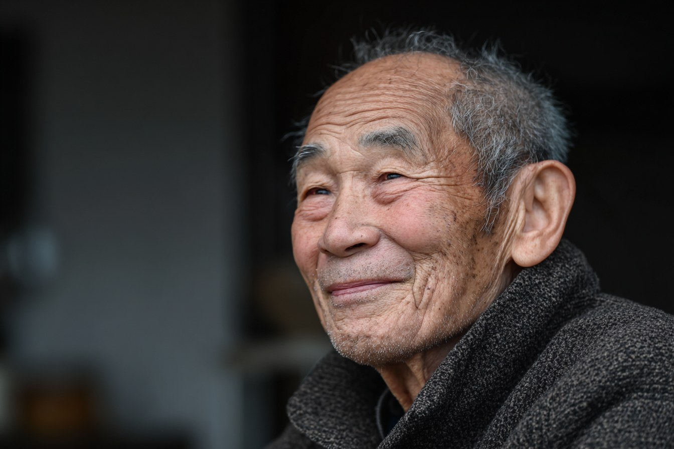 Retrato de um idoso sorridente, com pele enrugada e cabelo grisalho, vestindo um casaco escuro. A imagem transmite calor humano e sabedoria, capturada em um ambiente suave e iluminado.