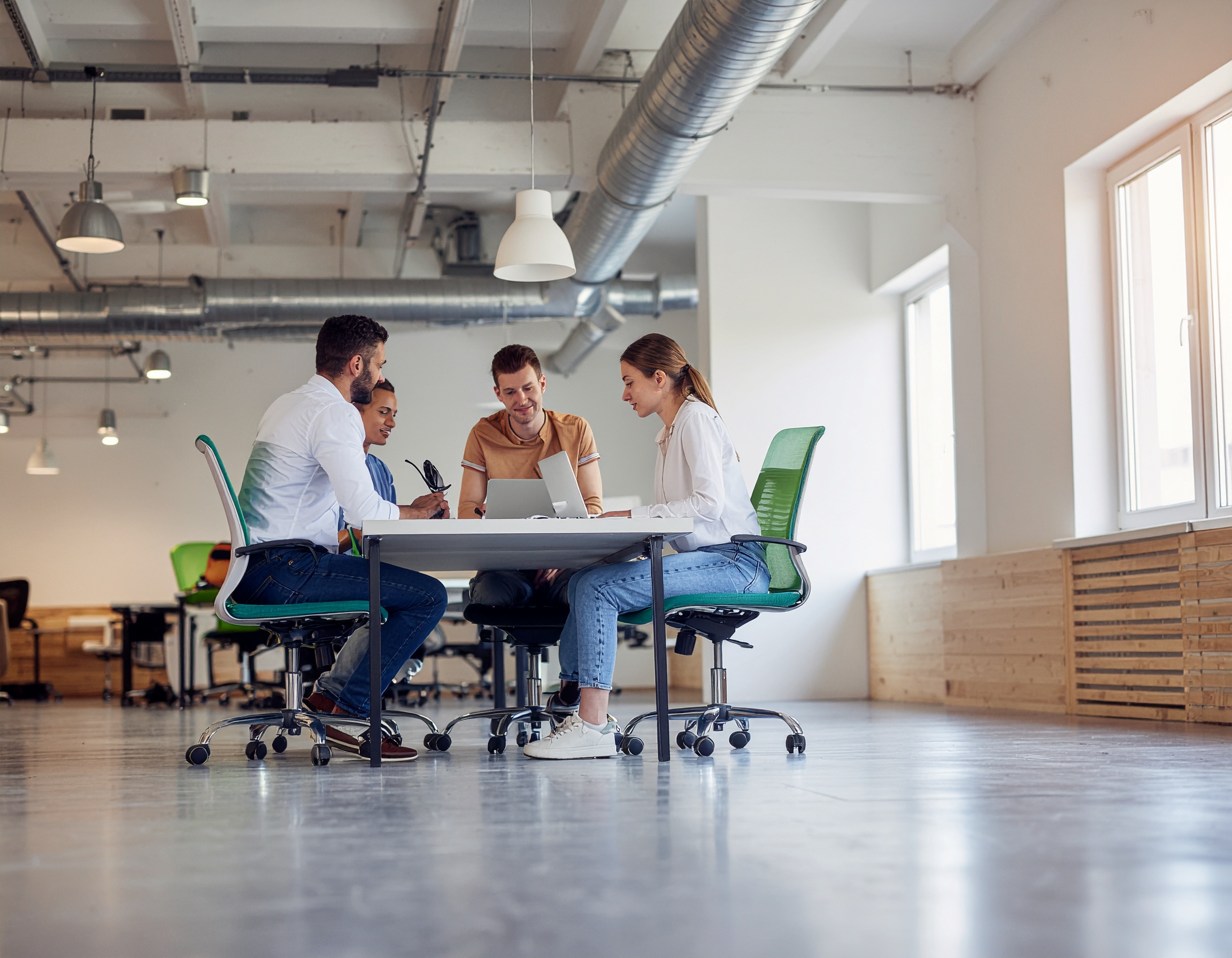 Grupo de quatro pessoas em um escritório moderno, sentadas ao redor de uma mesa discutindo em frente a um laptop. A iluminação natural entra pelas janelas amplas, criando um ambiente claro e colaborativo. O espaço é amplo e minimalista com cadeiras verdes e elementos metálicos visíveis no teto, refletindo um estilo industrial e contemporâneo.