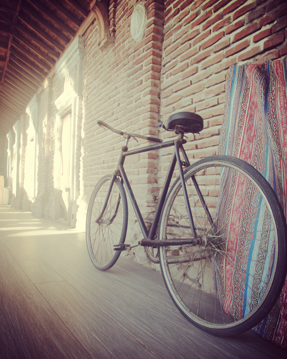 Bicicleta vintage encostada em parede de tijolos expostos, cercada por luz suave filtrando através de um corredor coberto. Tapete colorido adiciona textura vibrante ao ambiente rústico, criando uma composição equilibrada e nostálgica.