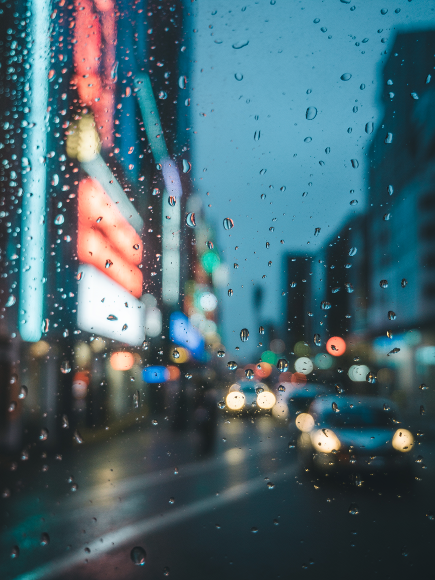 Cena urbana noturna vista através de uma janela com gotas de chuva, onde luzes de néon coloridas e faróis de carros criam um efeito bokeh, sugerindo uma atmosfera vibrante e movimentada. O foco nas gotas de água adiciona textura, enquanto o fundo desfocado realça a profundidade da imagem, capturando a essência dinâmica de uma cidade sob a chuva.