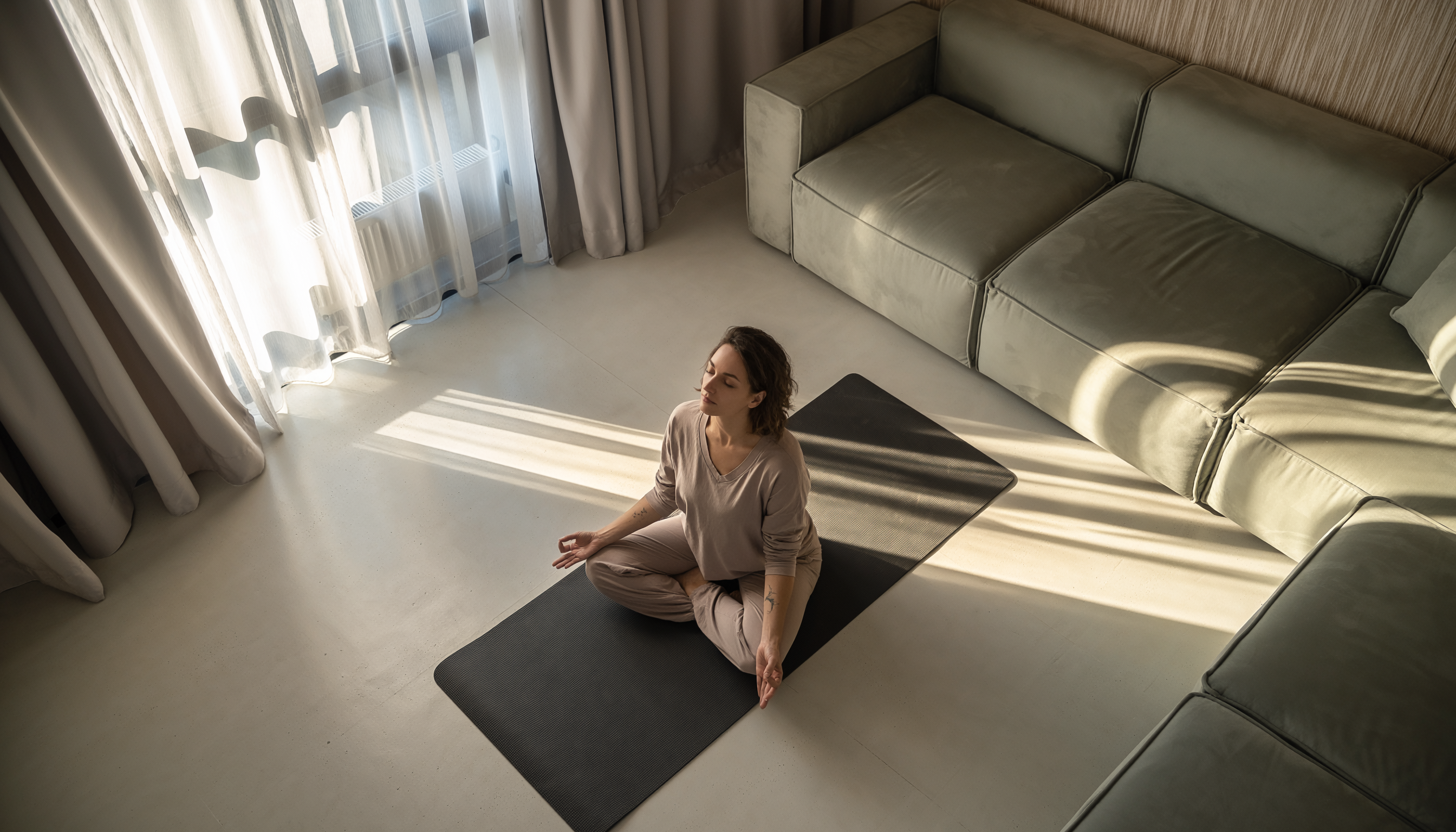 Mulher meditando em um tapete de ioga em uma sala bem iluminada, com sofás ao redor e cortinas permitindo a entrada suave da luz do sol. A composição é minimalista e serena, com tons neutros e atmosfera tranquila. A perspectiva de cima captura a cena em um ângulo aberto, destacando a harmonia entre a iluminação natural e o espaço organizado.