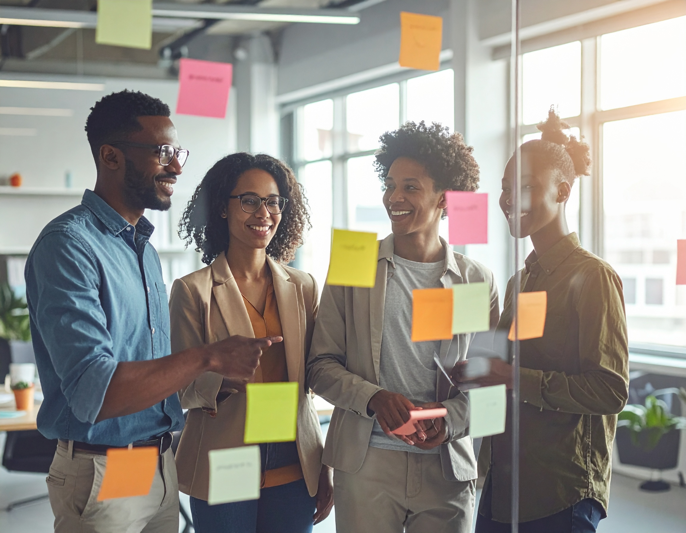 Quatro pessoas em uma reunião de equipe, discutindo ideias em um escritório moderno. Elas estão cercadas por notas adesivas coloridas em um painel de vidro. A iluminação natural atravessa as janelas amplas, criando uma atmosfera colaborativa e criativa. A cena é capturada em um ângulo ligeiramente inclinado, com foco claro nos rostos sorridentes e na interação entre os membros da equipe.