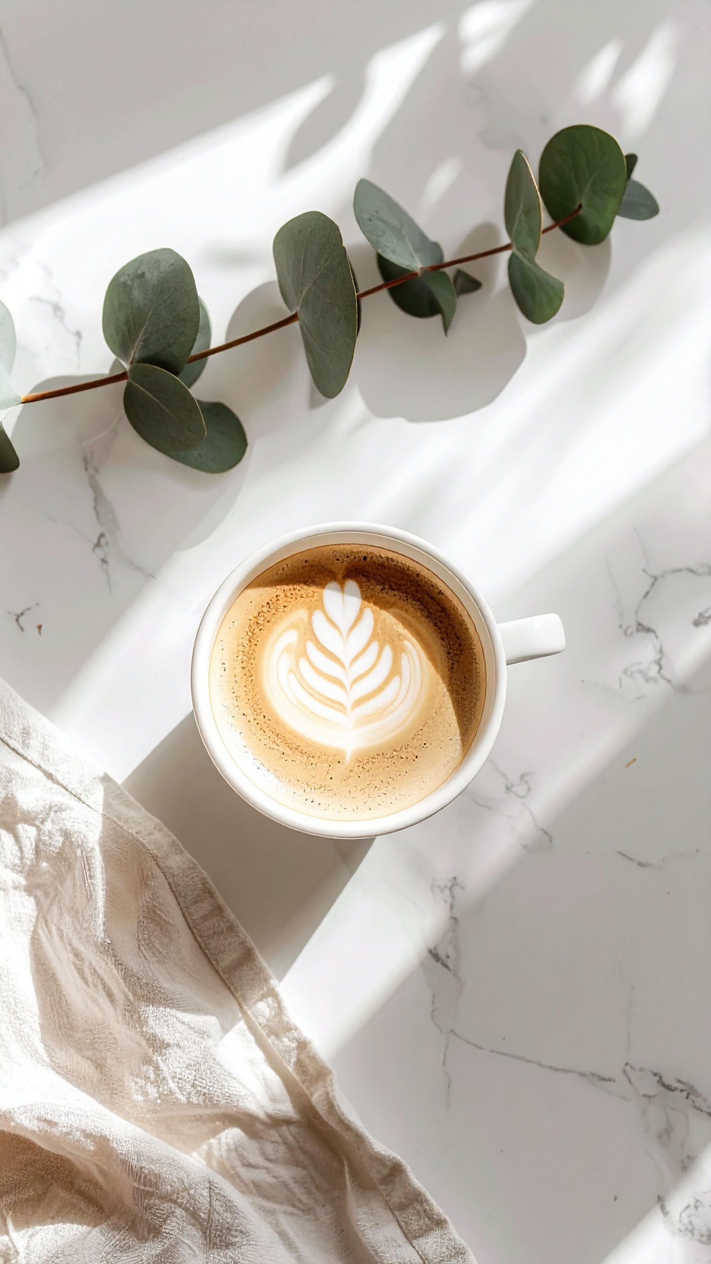 A latte art coffee sits in a white cup on a marble surface beside eucalyptus leaves