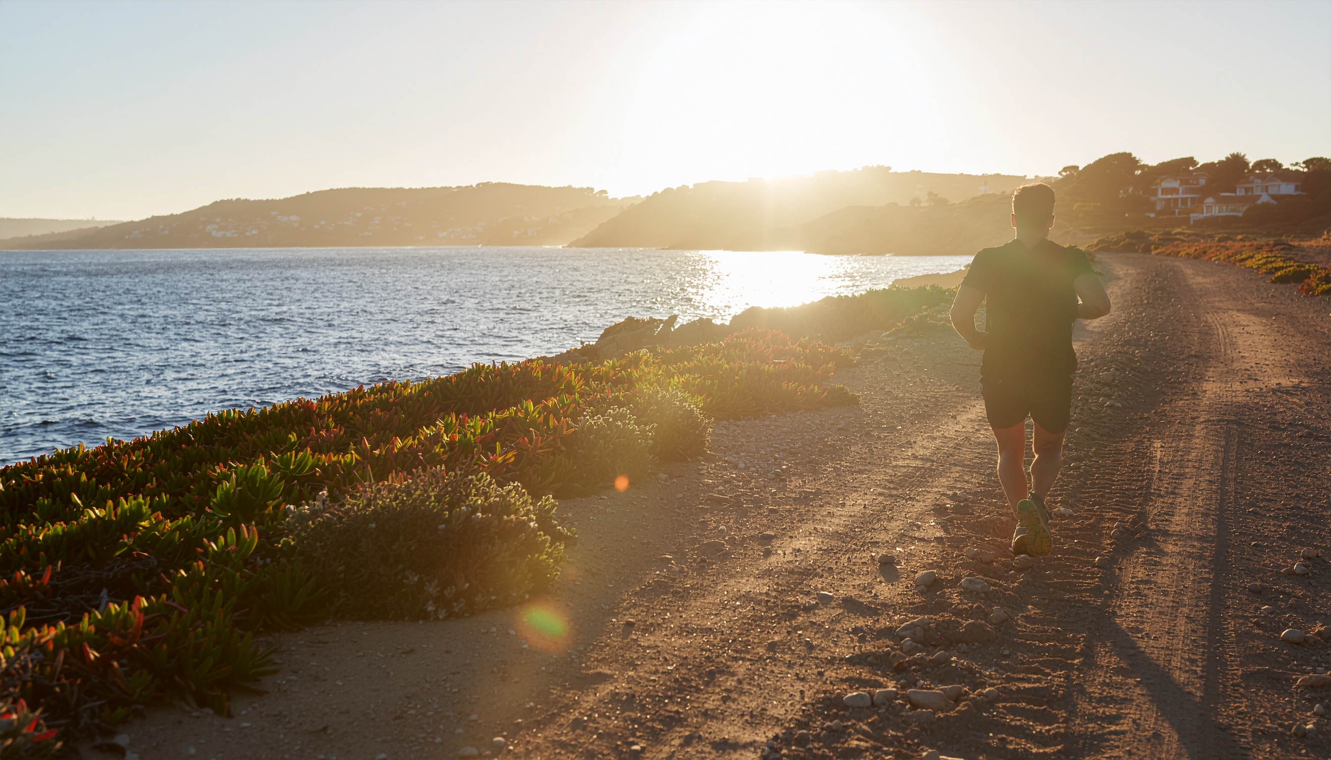 Homem correndo em uma trilha costeira ao pôr do sol, com vegetação rasteira e um vasto oceano à esquerda. A iluminação dourada do sol cria uma atmosfera serena e a composição destaca o contraste entre o caminho rústico e a paisagem natural. O ângulo da imagem enfatiza a perspectiva do corredor, enquanto o foco suave captura a beleza natural do cenário.