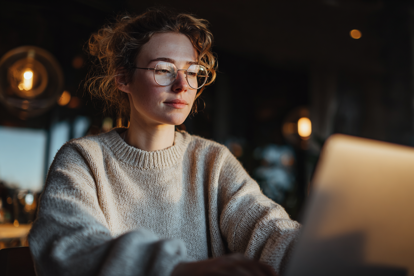 Pessoa concentrada em tela de laptop em ambiente interno com iluminação suave e quente. Usa suéter bege e óculos, com expressão de foco. Luzes desfocadas ao fundo criam atmosfera acolhedora. Composição centrada e natural, destacando características faciais sob luz dourada do entardecer.