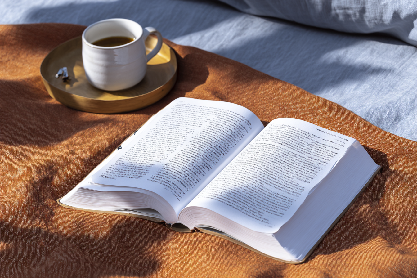 Livro aberto em tecido laranja, com xícara de café sobre bandeja dourada ao lado. Iluminação suave e sombras criam atmosfera acolhedora, destacando textura do tecido. Composição harmônica e relaxante.