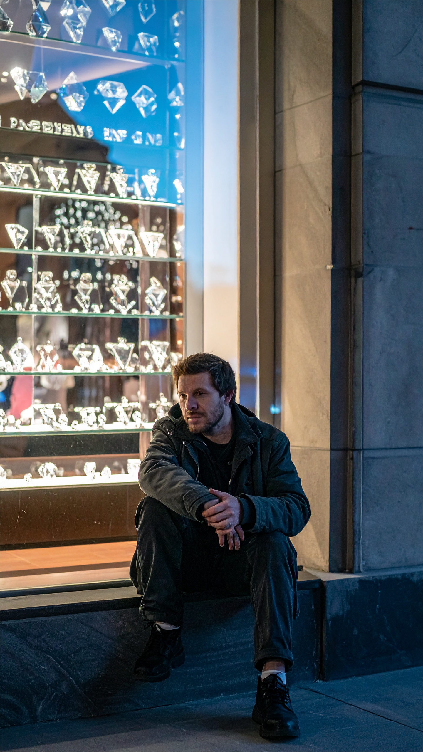 A man sits pensively outside a brightly lit jewelry store window, showcasing numerous sparkling diam