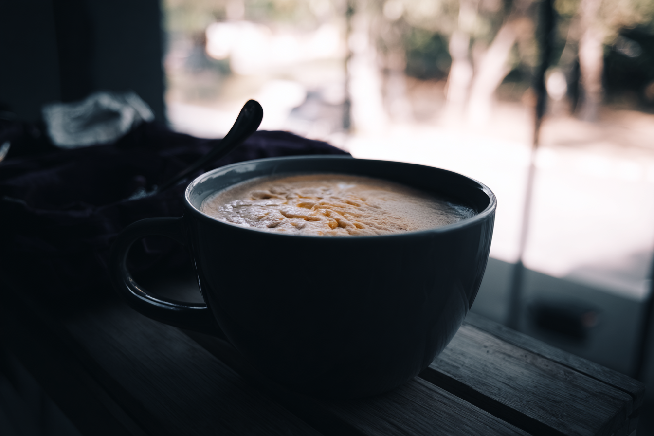 Uma xícara de café espumante sobre uma superfície de madeira em um ambiente com luz natural suave, fundos desfocados, transmitindo um clima acolhedor e tranquilo. A composição é simples, com foco nítido no café, destacando a textura cremosa da espuma sob luz natural que entra pela janela.