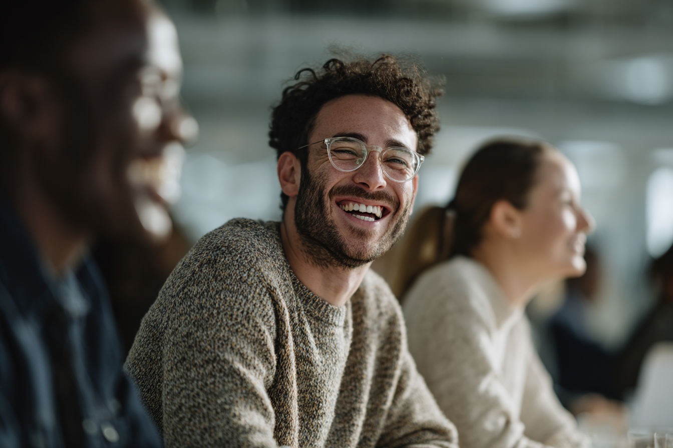 Um grupo de jovens sorridentes em um ambiente de trabalho moderno, destacando um homem de cabelo cacheado e óculos, que demonstra alegria e camaradagem. A imagem captura a essência da colaboração e interação social em um escritório.