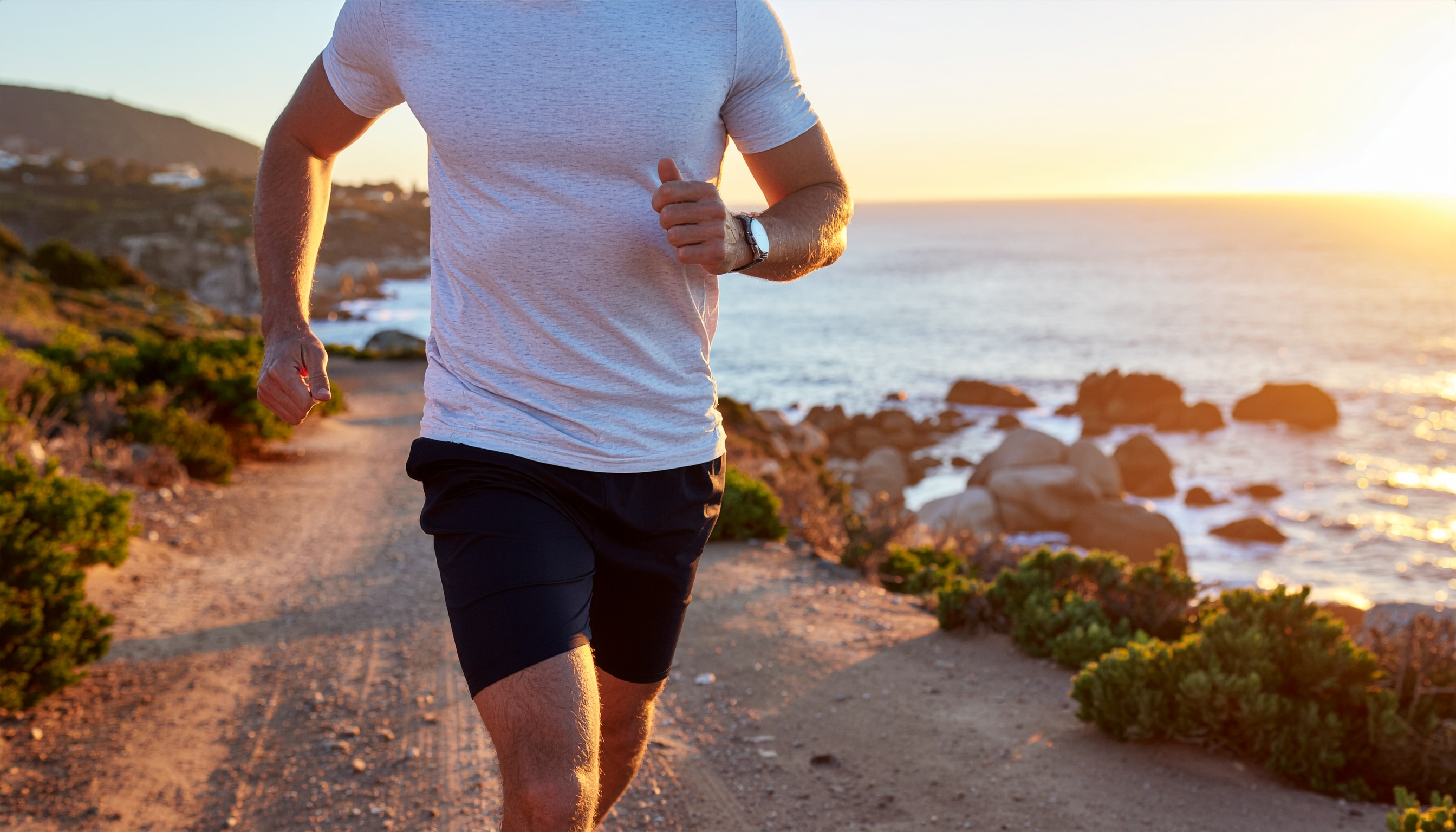 Homem correndo ao longo de uma trilha costeira durante o pôr do sol, vestindo camiseta branca e shorts escuros, com uma vista deslumbrante do oceano ao fundo. A luz dourada do sol ilumina o cenário, destacando os contornos do corpo do corredor e as formações rochosas à beira-mar. A composição captura a essência de um estilo de vida ativo e saudável em harmonia com a natureza.