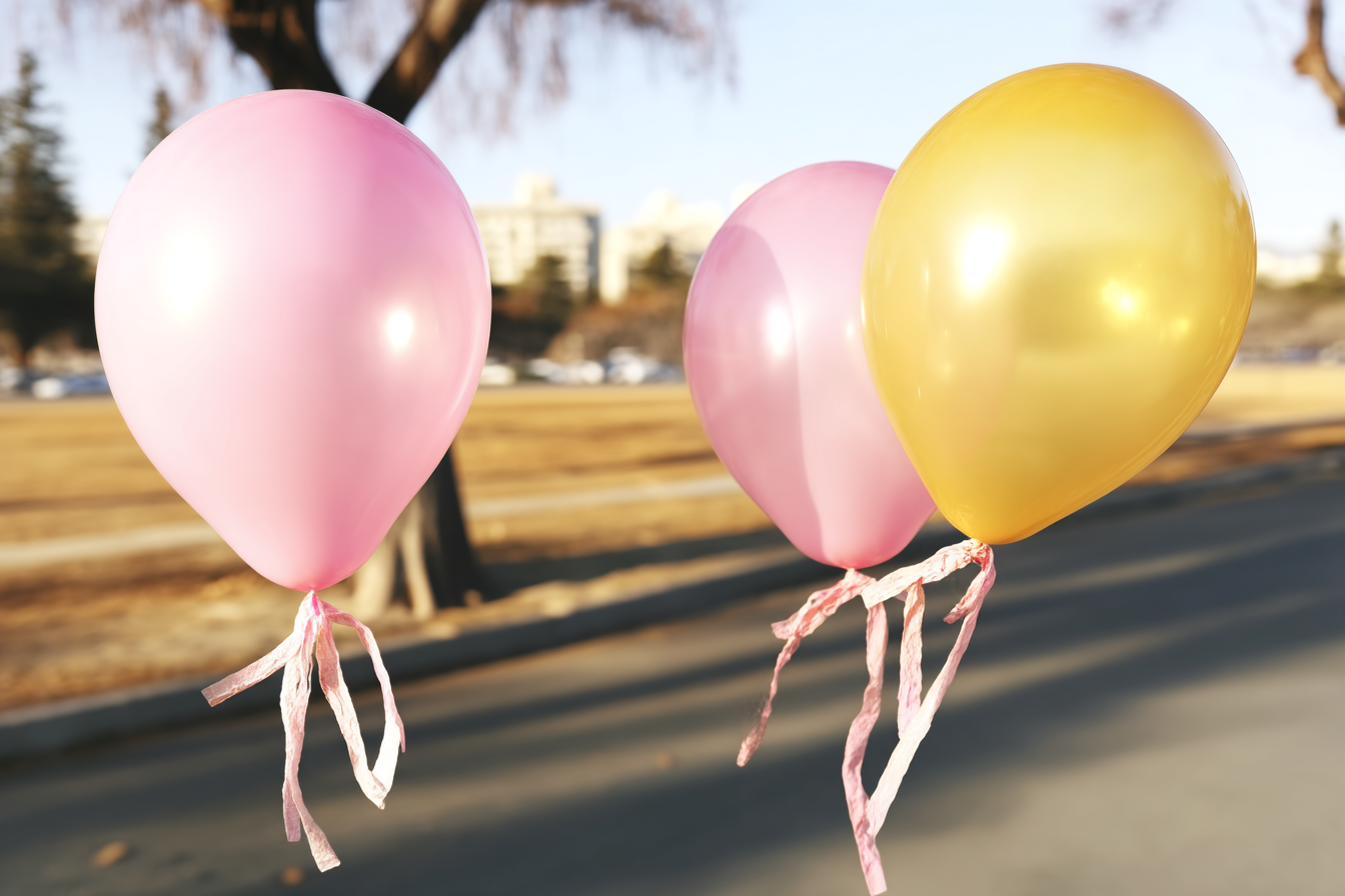 A imagem apresenta três balões coloridos flutuando, dois na tonalidade rosa e um na tonalidade dourada. Os balões têm um brilho suave e refletem a luz do sol, destacando suas superfícies lisas e brilhantes. As fitas que os prendem ao chão são de cor rosa claro e têm um aspecto esvoaçante. Ao fundo, um cenário de parque pode ser visto com árvores e um céu claro.