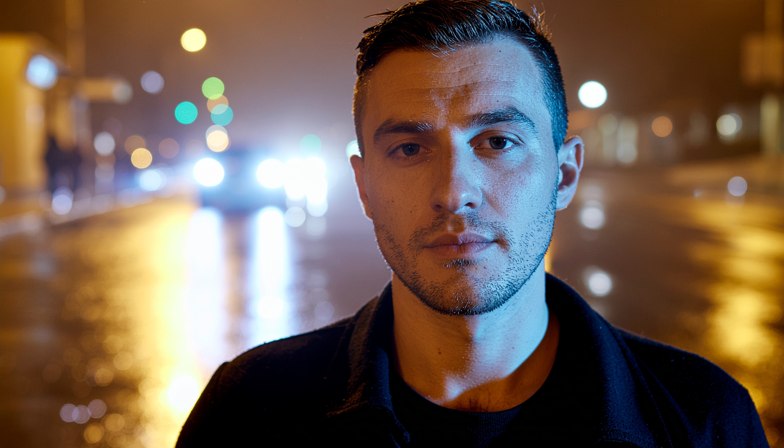 Retrato de um homem em uma rua à noite, com iluminação mista azul e amarela refletida na pele. O fundo desfocado mostra luzes de carros e postes, criando um efeito bokeh. A expressão séria e a iluminação dramática conferem uma atmosfera de introspecção e mistério.