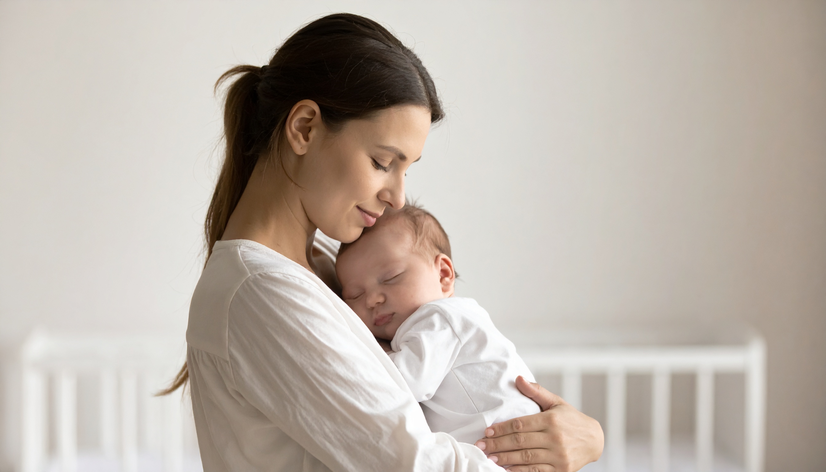 Mãe abraça carinhosamente seu bebê adormecido em ambiente sereno, com iluminação suave e tons neutros, transmitindo ternura e tranquilidade. A composição foca na conexão emocional, com fundo desfocado em um quarto infantil minimalista.