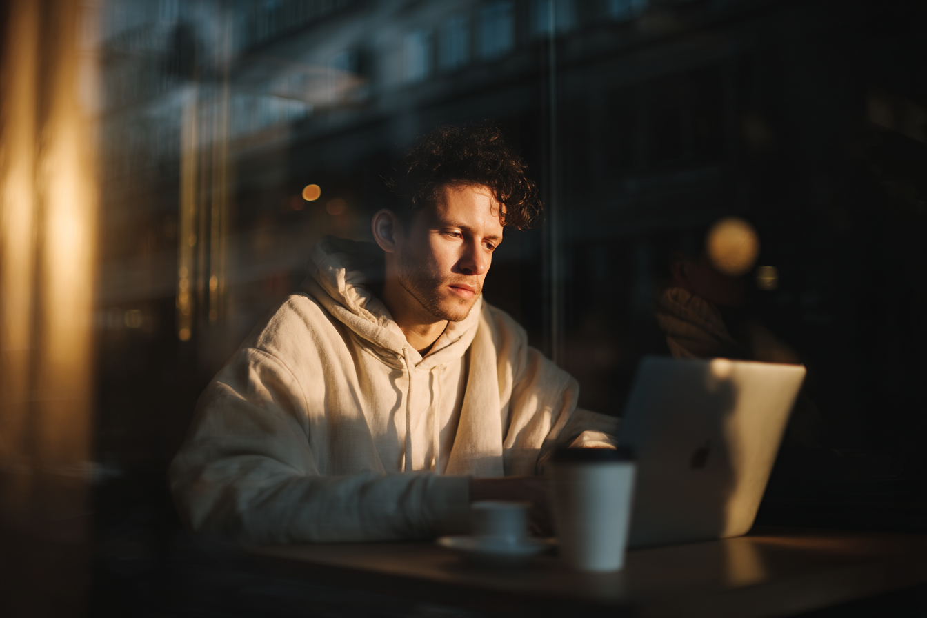 A imagem apresenta um jovem em um ambiente iluminado suavemente, focado em seu laptop. As cores predominantes incluem tons quentes de dourado e creme, com um fundo mais escuro que sugere um espaço urbano. A luz natural parece filtrada, criando um efeito de brilho ao redor do sujeito e dos objetos em primeiro plano. O jovem veste um moletom claro, e há um copo de café e um pires na mesa, ambos com tons neutros.