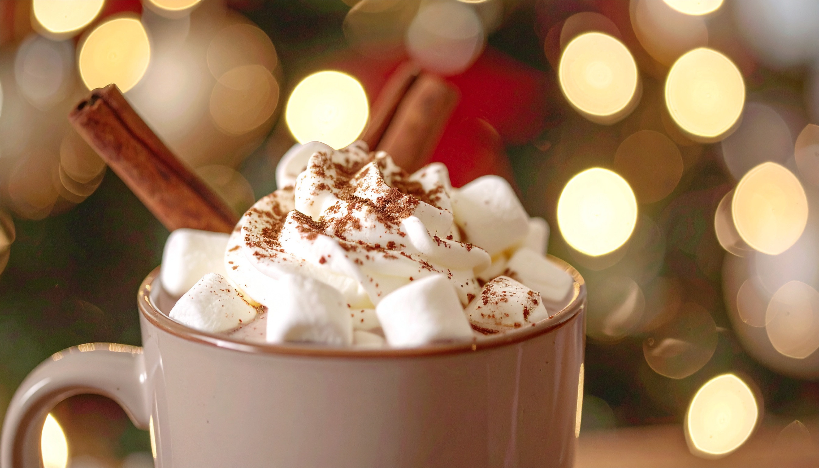 Caneca de chocolate quente com chantilly e marshmallows, decorada com canela em pau. A iluminação suave cria um bokeh festivo ao fundo, acentuando uma atmosfera acolhedora e natalina.