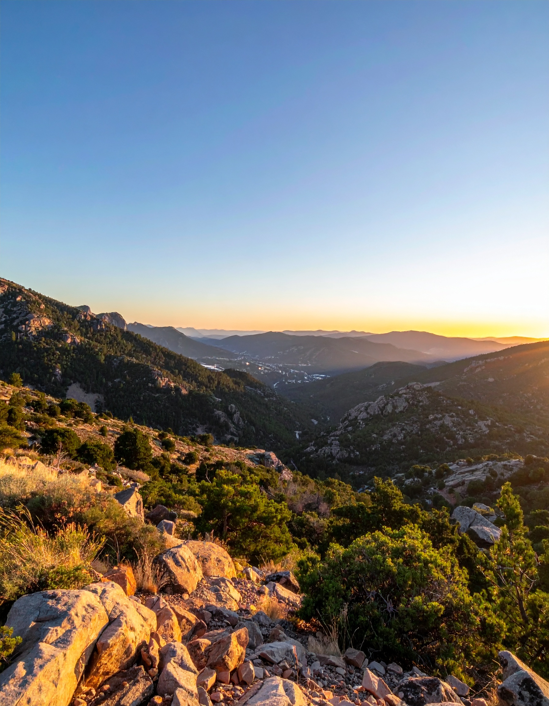 Vista panorâmica de montanhas ao entardecer, com vegetação densa e rochas sob luz dourada. O céu claro e azul se encontra com o horizonte, criando uma atmosfera serena e ampla. A composição é equilibrada, com profundidade destacada por sombras suaves e foco nítido nas texturas naturais.