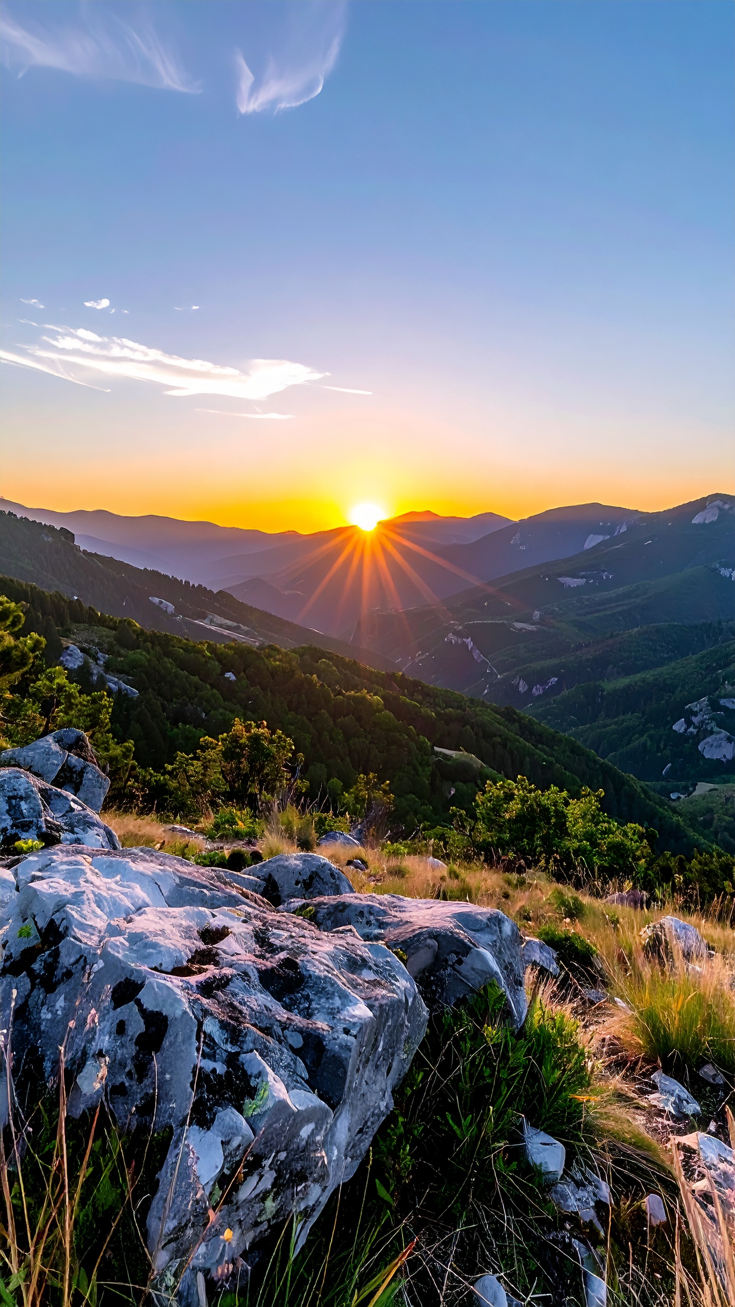 Paisagem de montanha ao pôr do sol, com luz dourada iluminando o horizonte e criando sombras longas sobre rochas e vegetação. Céu azul claro com nuvens delicadas e composição equilibrada, transmitindo tranquilidade e beleza natural.