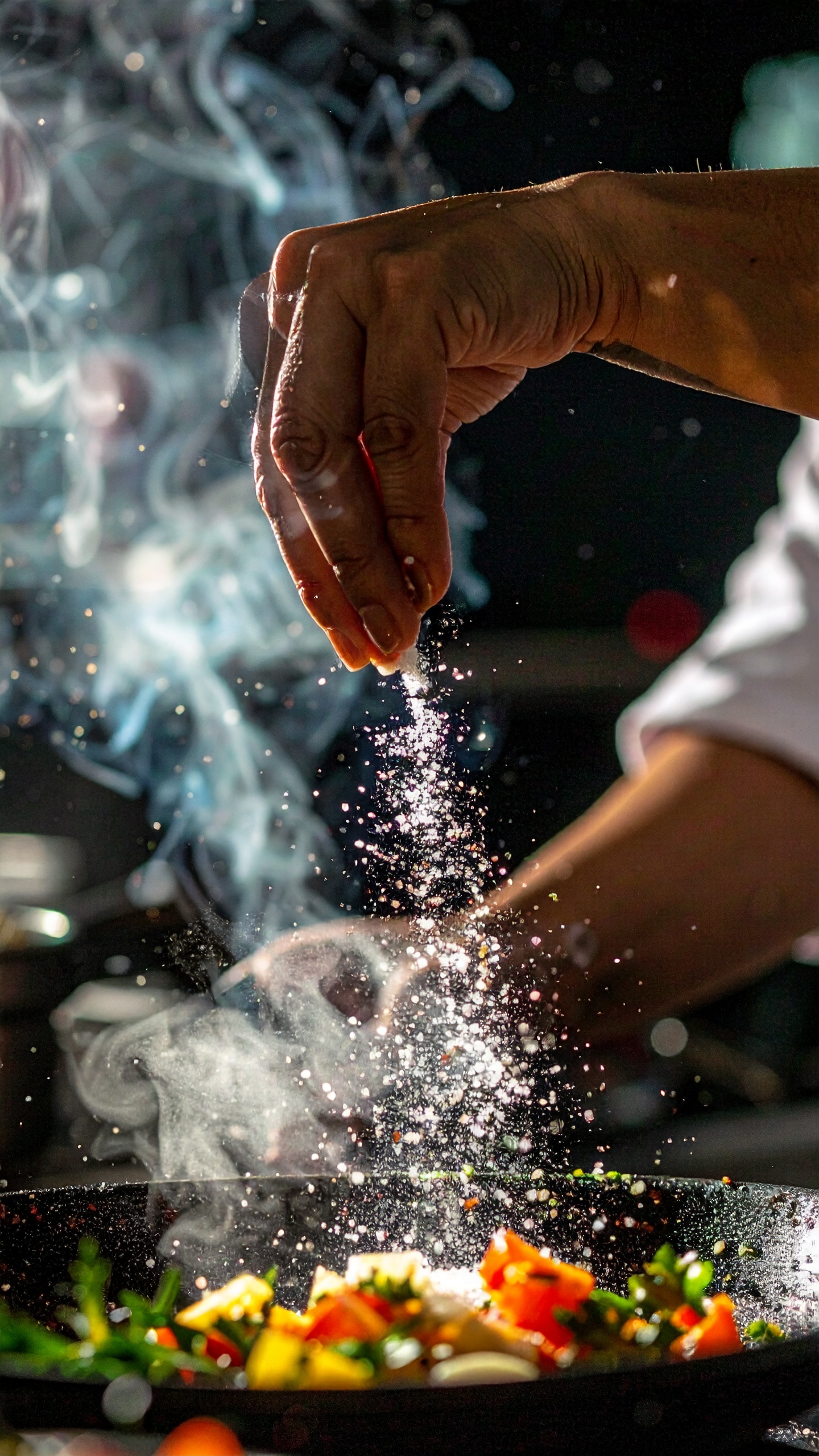 A hand sprinkles salt over a sizzling pan filled with colorful vegetables