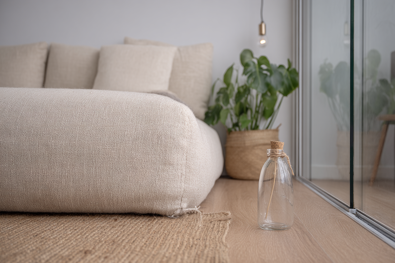 Sala de estar minimalista com sofá bege de tecido, planta em vaso de fibras naturais e garrafa de vidro com tampa de cortiça em piso de madeira. A composição é iluminada suavemente por lâmpada pendente, criando uma atmosfera serena e acolhedora. A luz natural entra pela porta de vidro à direita, realçando as texturas e as cores neutras.