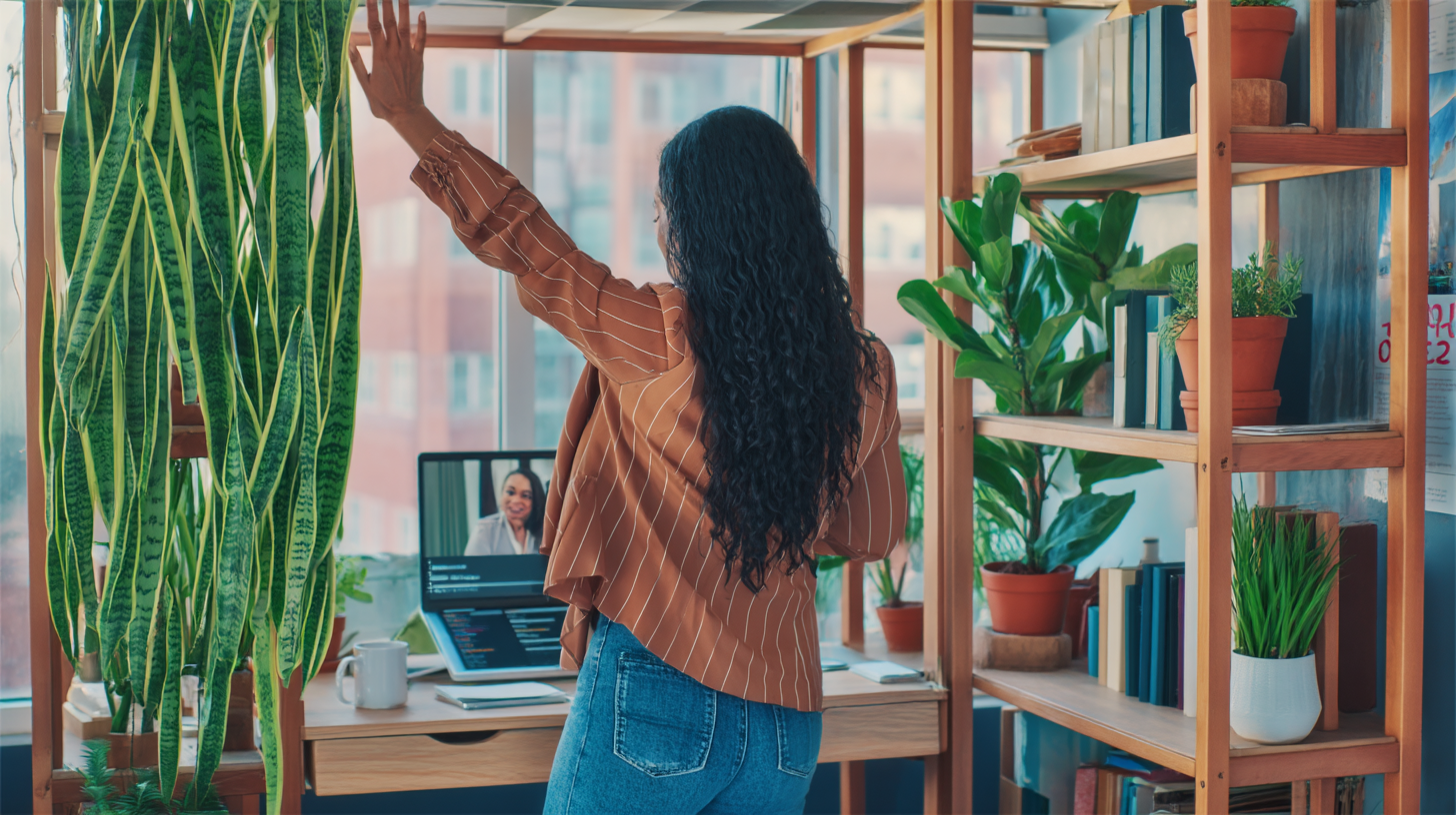 Mulher em home office acena para tela durante videoconferência, cercada por plantas e estantes organizadas, transmitindo produtividade e conexão em ambiente doméstico acolhedor.