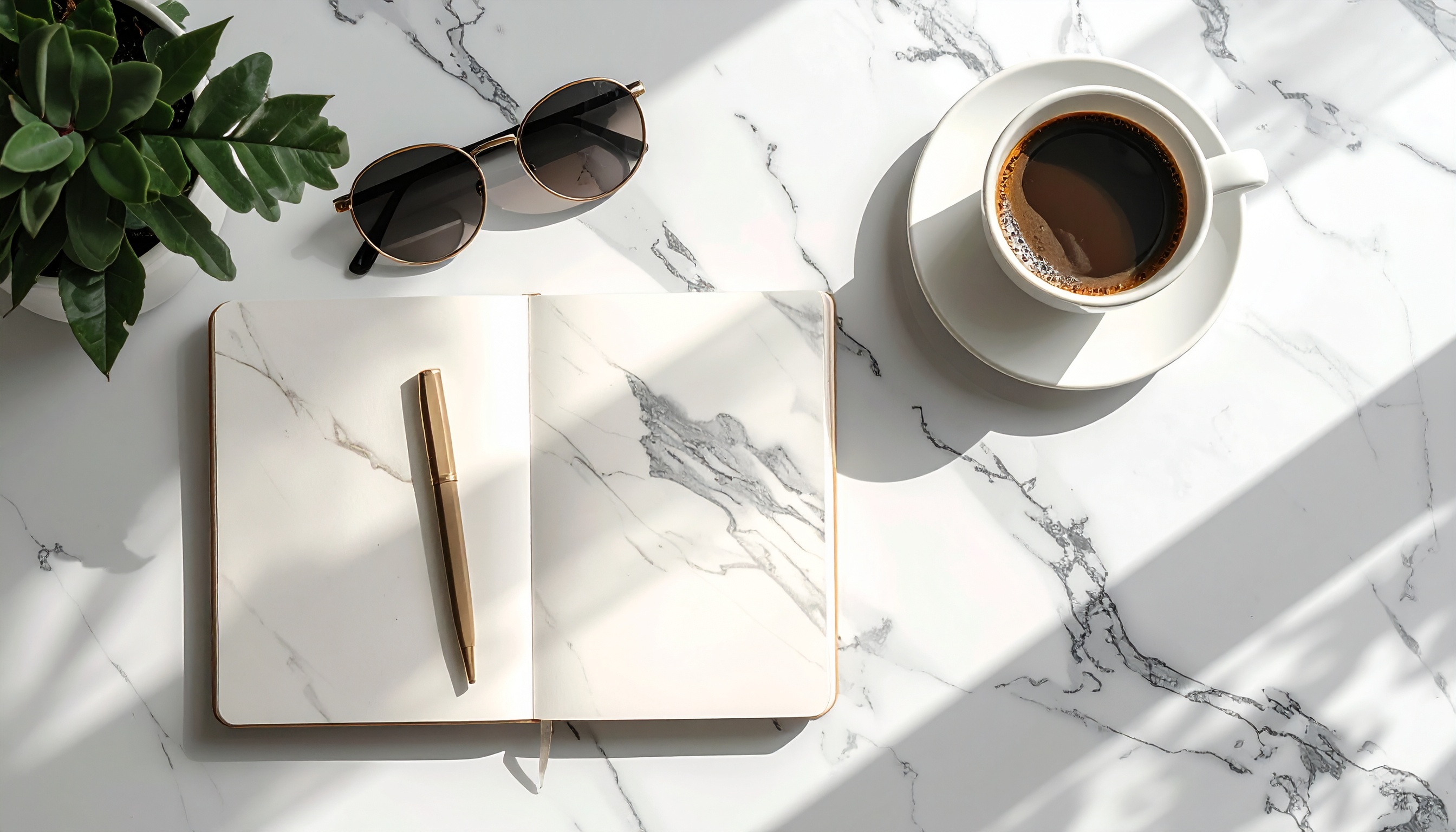 Cena minimalista com caderno aberto de capa marmorizada, caneta dourada ao lado de óculos de sol, xícara de café e planta em fundo de mármore claro. Luz suave e sombras projetadas criam um ambiente elegante e tranquilo.