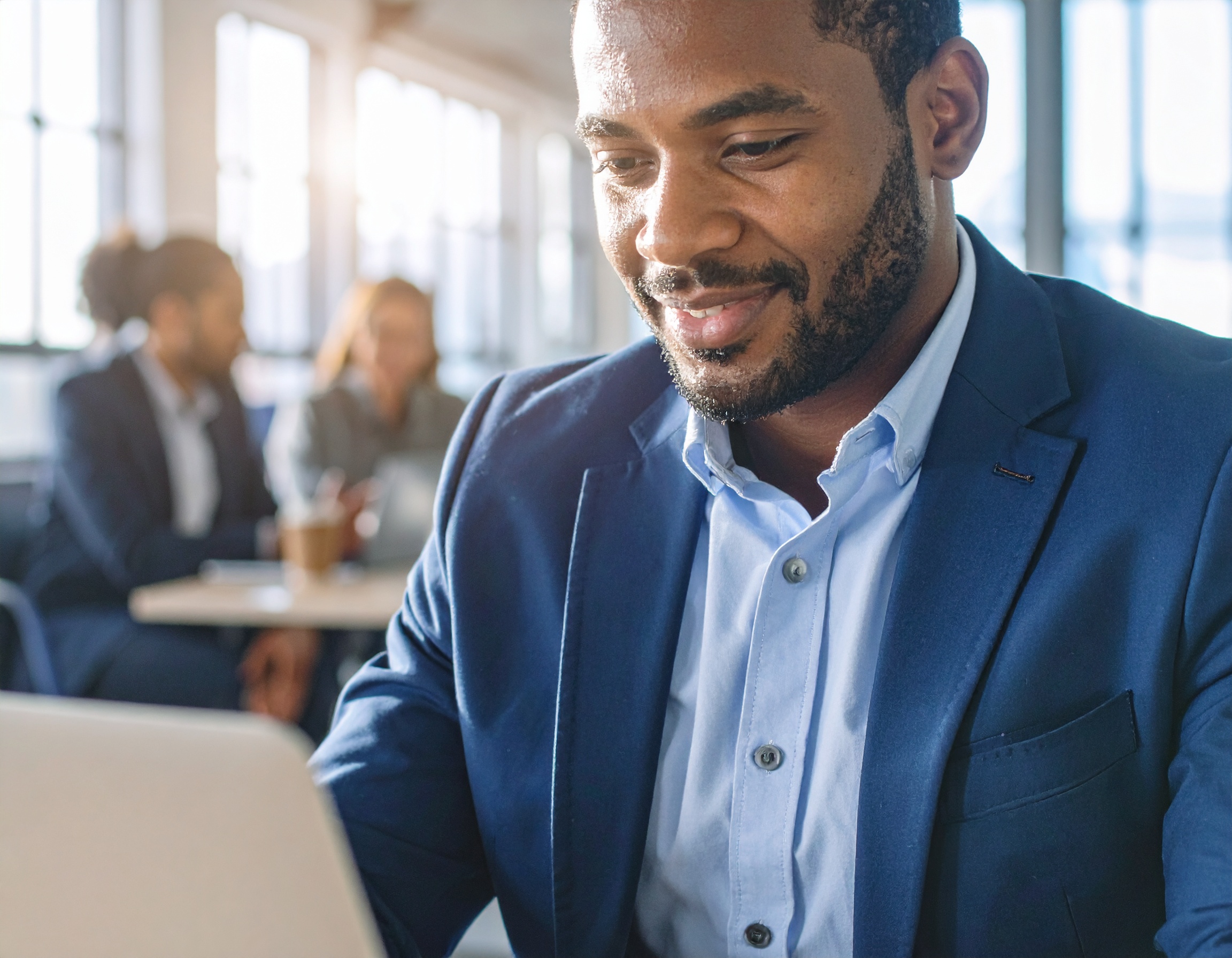 Homem sorridente usando terno azul, focado em laptop em ambiente de escritório moderno com iluminação natural suave. Pessoas ao fundo desfocadas, criando atmosfera colaborativa e profissional. Composição equilibrada com foco nítido no rosto do homem, transmitindo concentração e dinamismo.