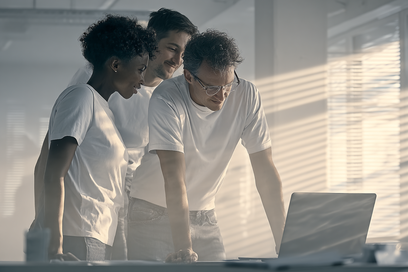 Três pessoas em pé ao redor de um laptop em um ambiente de escritório iluminado por luz natural suave, vestindo camisetas brancas. O foco está em sua colaboração e concentração, com sombras de persianas criando um padrão no fundo. A imagem transmite um ambiente de trabalho colaborativo e moderno, com uma composição equilibrada e iluminação natural acentuando os detalhes.