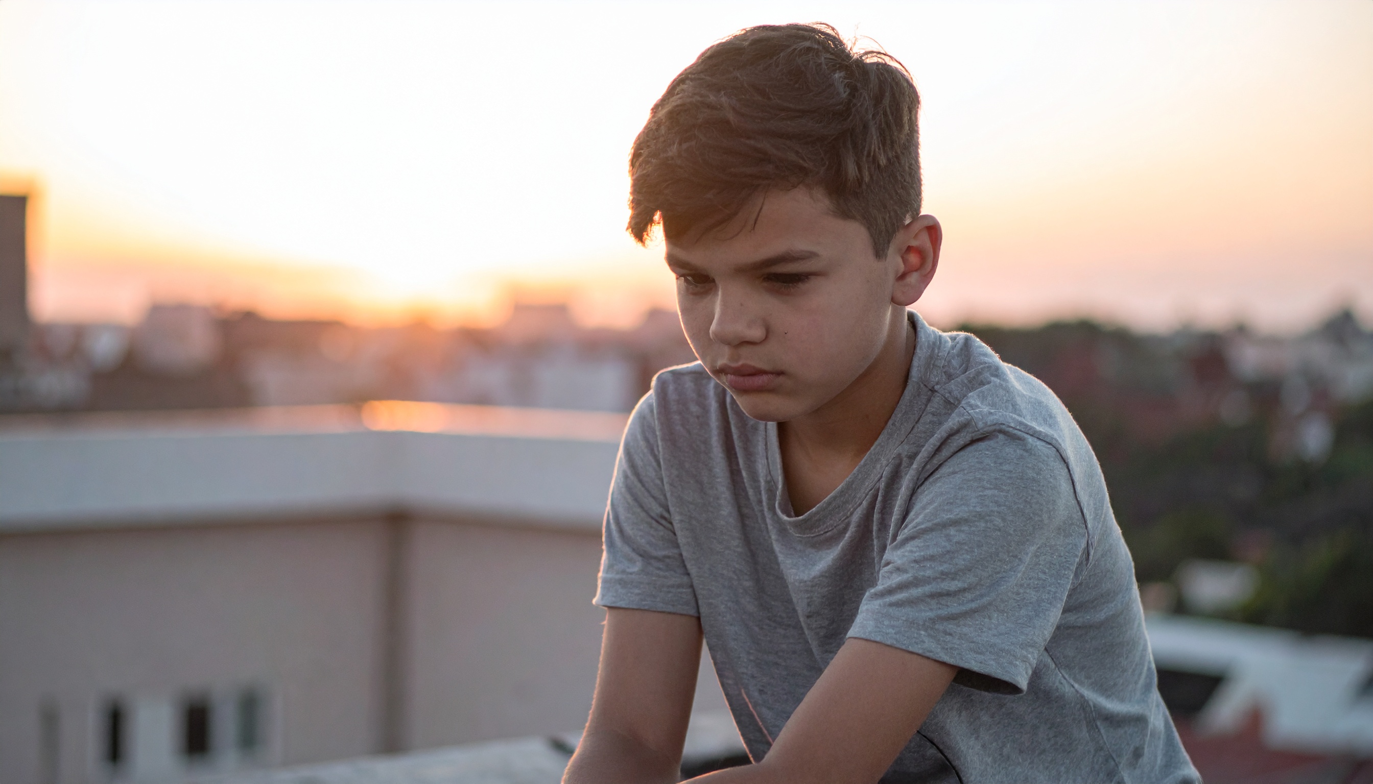 A young boy in a gray t-shirt sits thoughtfully on a rooftop during sunset