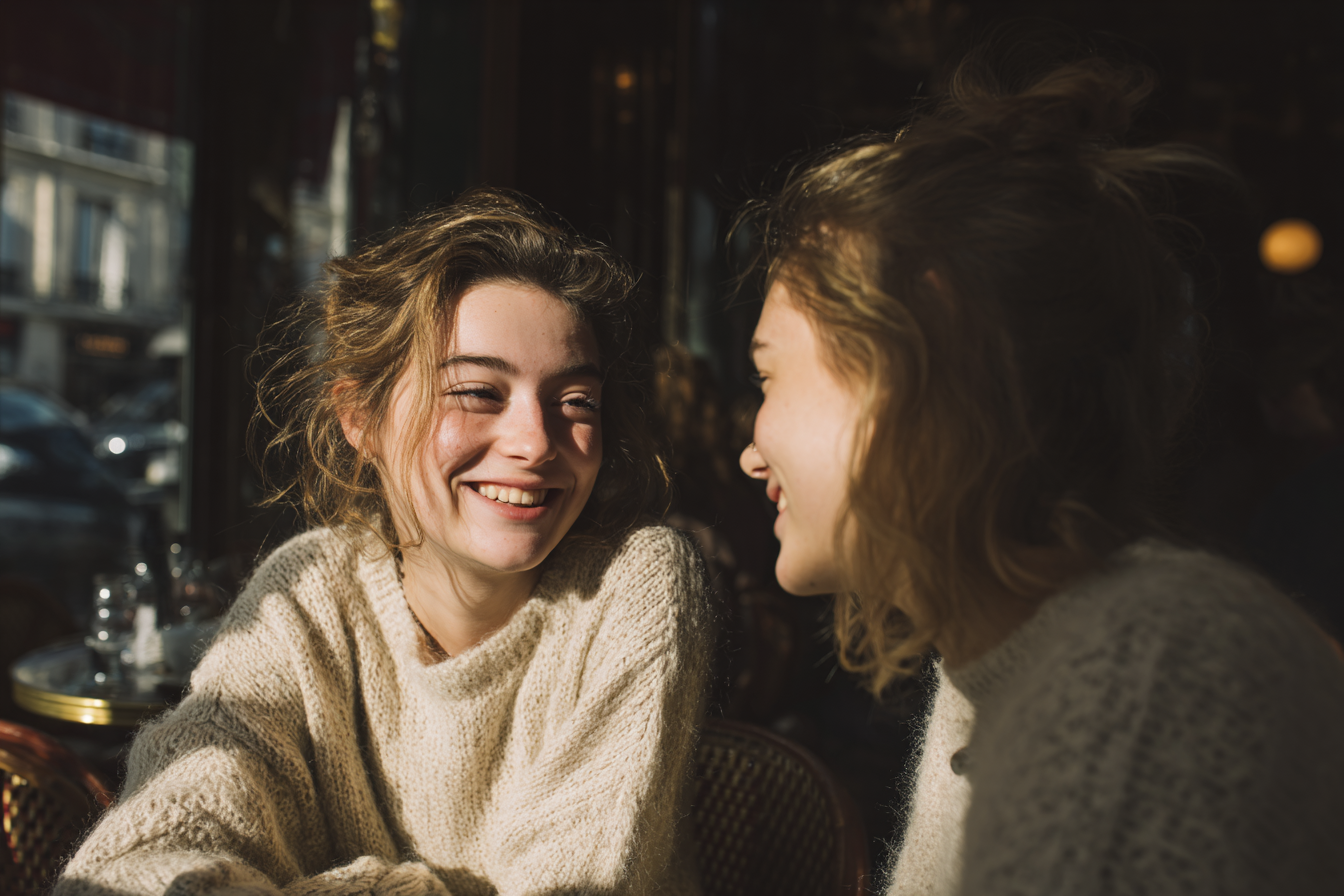 A imagem apresenta duas jovens sorrindo uma para a outra, capturadas em um ambiente iluminado por luz natural. As cores predominantes são tons quentes, com destaque para os beges e marrons dos suéteres, contrastando com um fundo mais escuro. A luz suave destaca os rostos das jovens, criando um efeito acolhedor. A composição é centrada nas duas figuras, com um fundo desfocado que sugere um café ou bistrô.