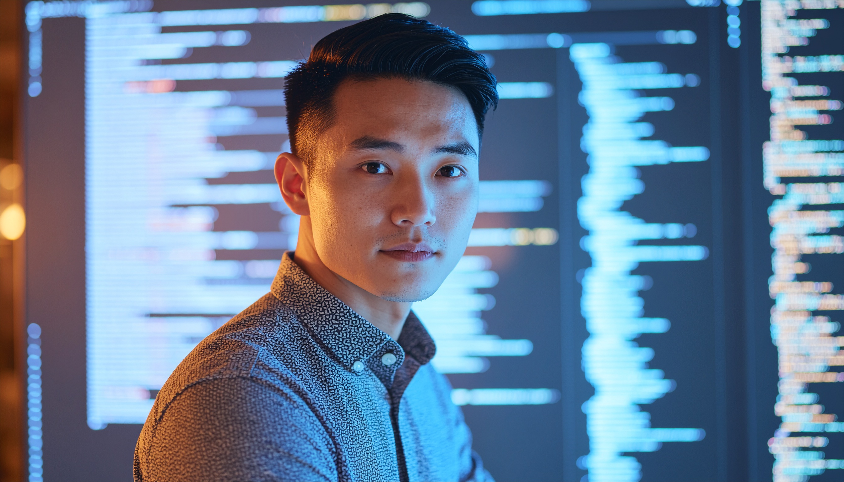 Um homem em um ambiente de alta tecnologia com telas de código ao fundo, destacando suas feições sob iluminação azulada. A composição enfatiza o profissionalismo e a concentração, com foco nítido na expressão do homem e no brilho dos monitores, criando uma atmosfera moderna e inovadora.