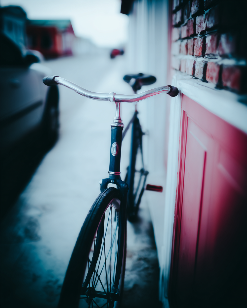 Bicicleta vintage encostada em parede de tijolos vermelhos, destacando-se em meio a um cenário urbano desfocado. A composição apresenta uma paleta de cores fria, contrastando com o vermelho da porta e dos tijolos. A iluminação suave e o foco seletivo criam uma atmosfera nostálgica e calma, com ênfase nos detalhes metálicos do guidão. O ângulo baixo da câmera permite uma perspectiva dinâmica da bicicleta e da rua adjacente.