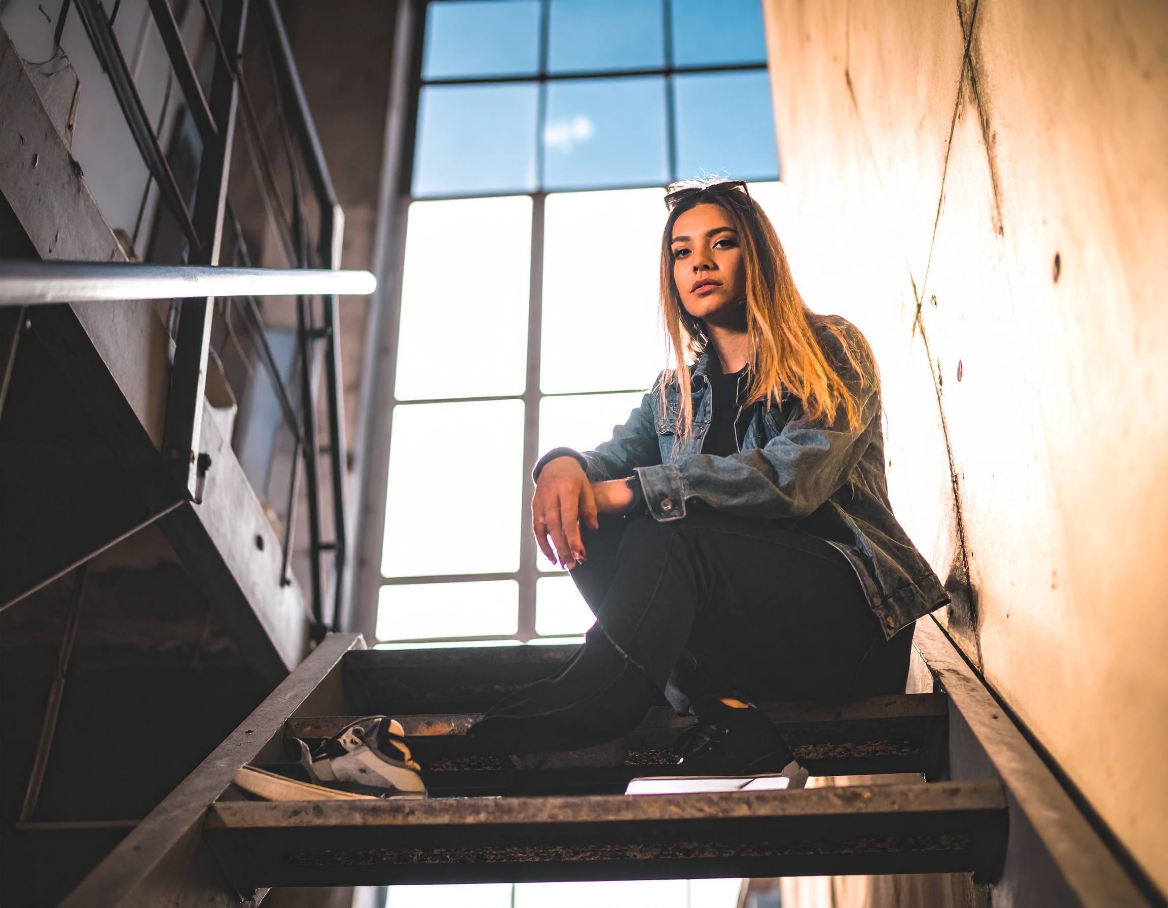 Jovem mulher sentada em escada industrial iluminada pela luz do sol, transmitindo confiança e estilo urbano moderno com jaqueta jeans e ambiente minimalista.