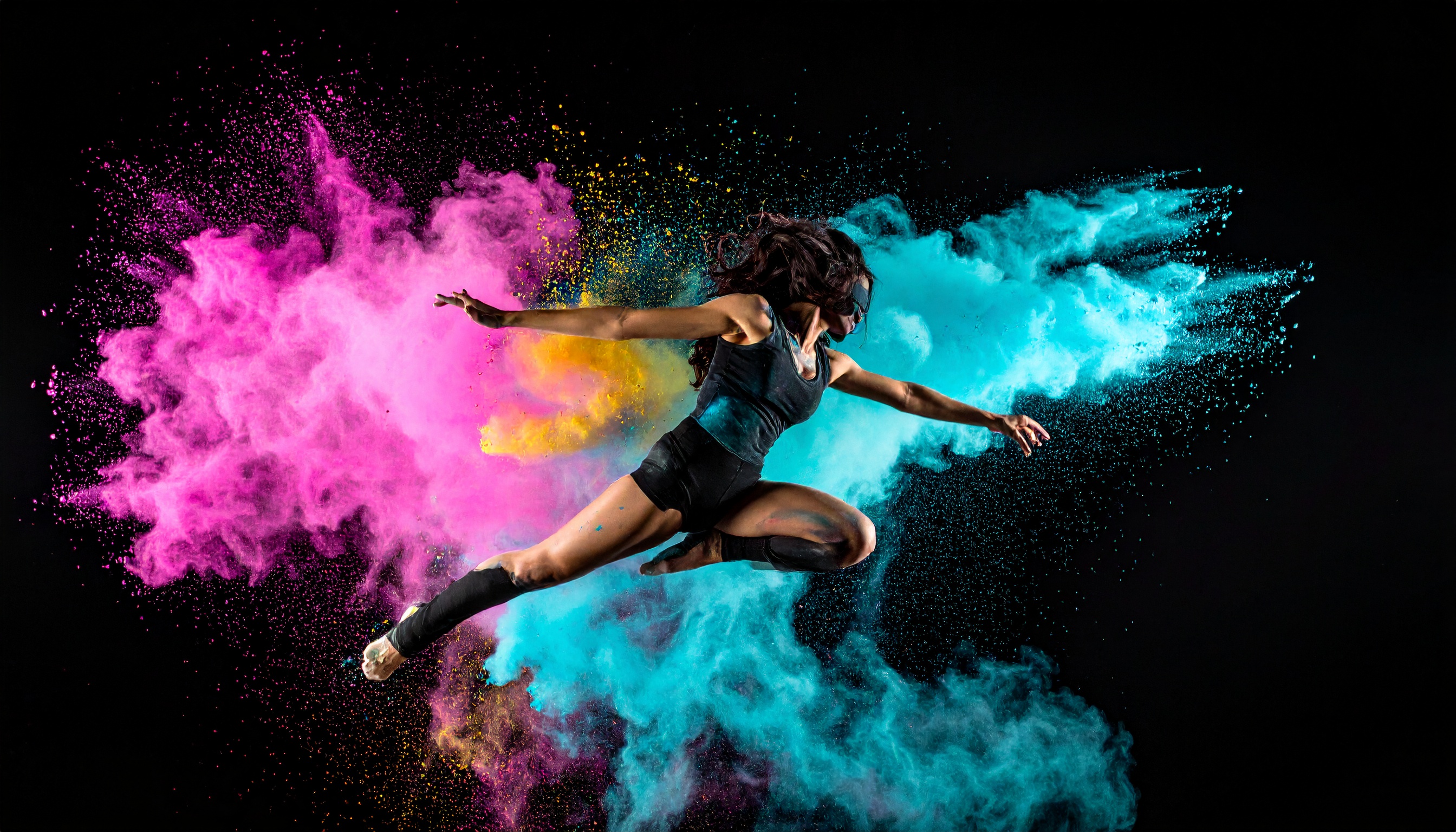 A dynamic dancer leaps through vibrant clouds of colored powder against a dark backdrop