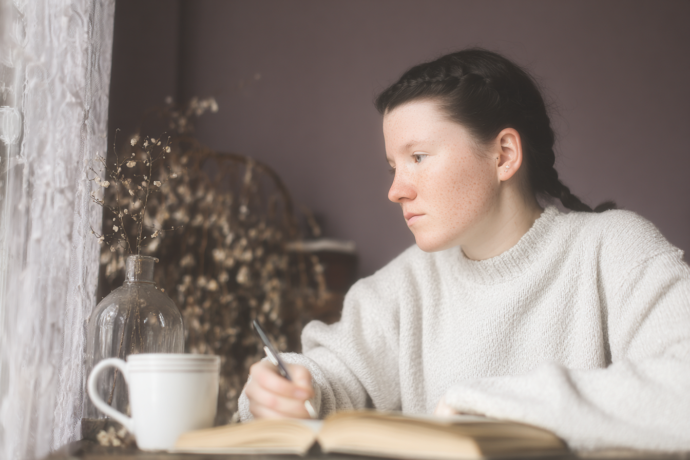 Jovem mulher sentada junto à janela, escrevendo em um caderno, com iluminação suave e delicada. A cena tem atmosfera tranquila, destacando uma blusa de lã, copo de café e vaso com flores secas. A composição harmoniosa e o foco suave criam um ambiente introspectivo e acolhedor.