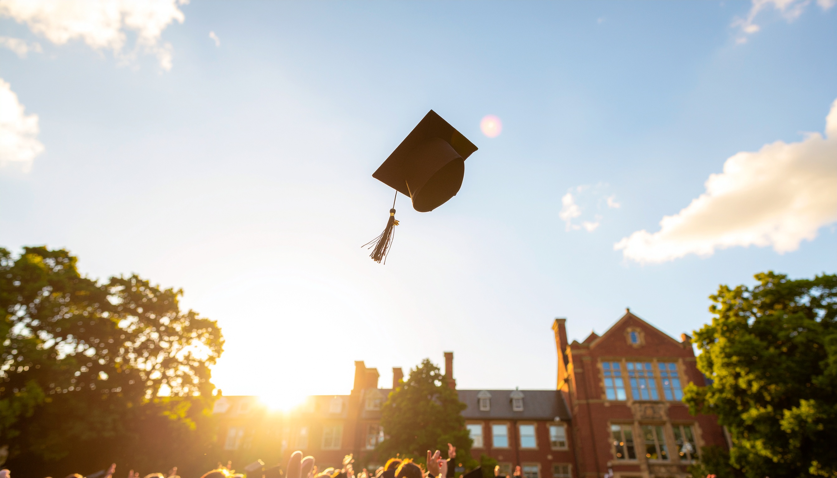 Um chapéu de formatura é lançado ao ar em um campus universitário durante a cerimônia de graduação ao pôr do sol, transmitindo alegria e realização.