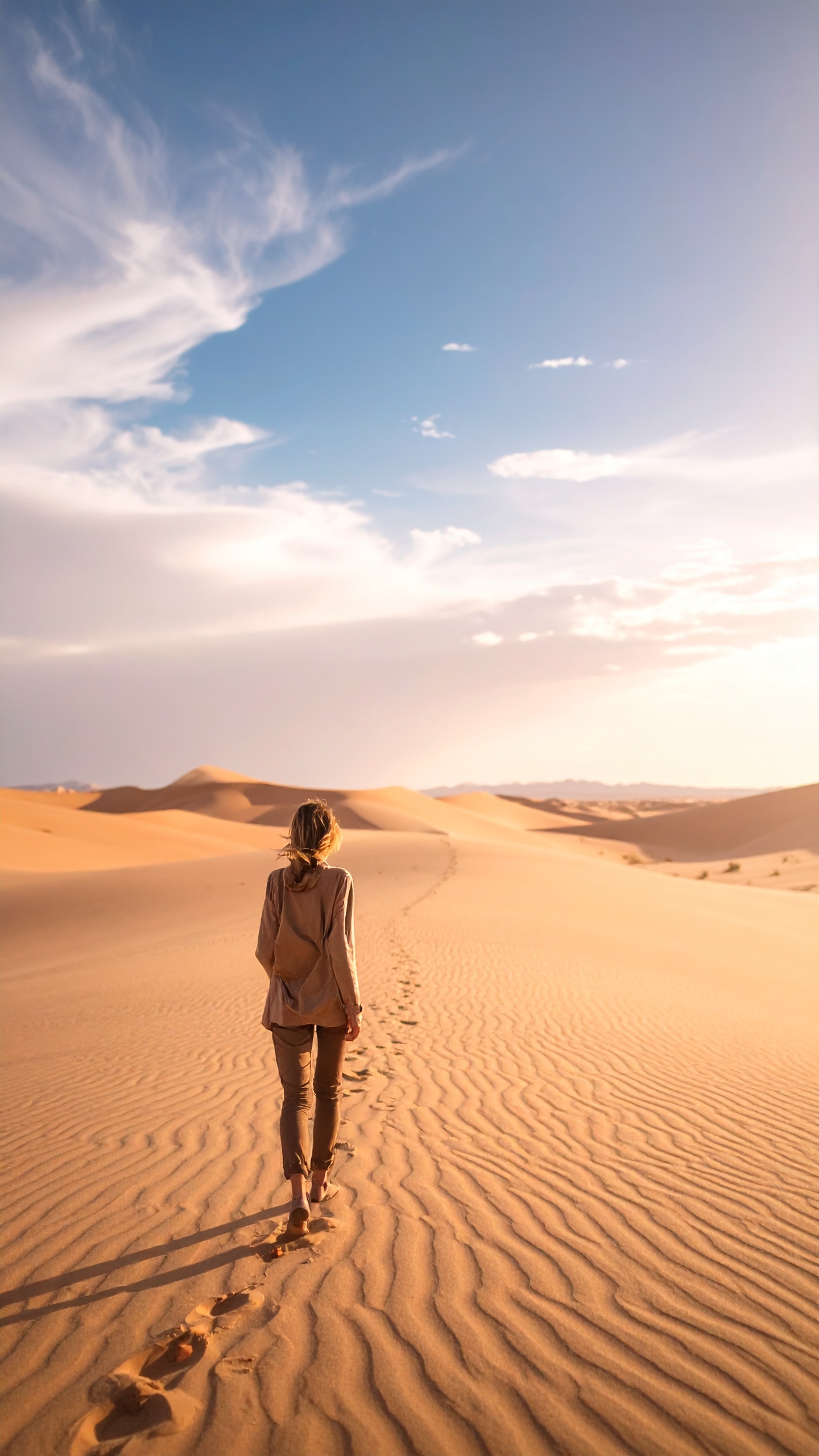 A lone traveler walks across sunlit desert dunes under a vast sky