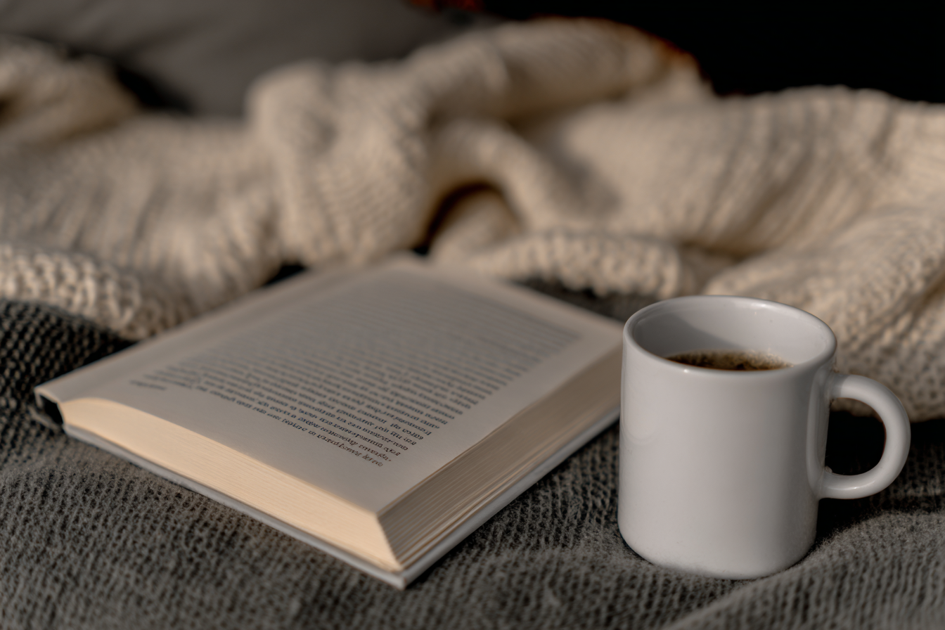 Cena aconchegante com um livro aberto ao lado de uma xícara de café em uma superfície coberta por tecidos tricotados. A iluminação suave e as cores neutras criam um ambiente de relaxamento perfeito para leitura em um dia frio. O foco está na xícara branca e nas páginas do livro, destacando o conforto e a tranquilidade da cena.